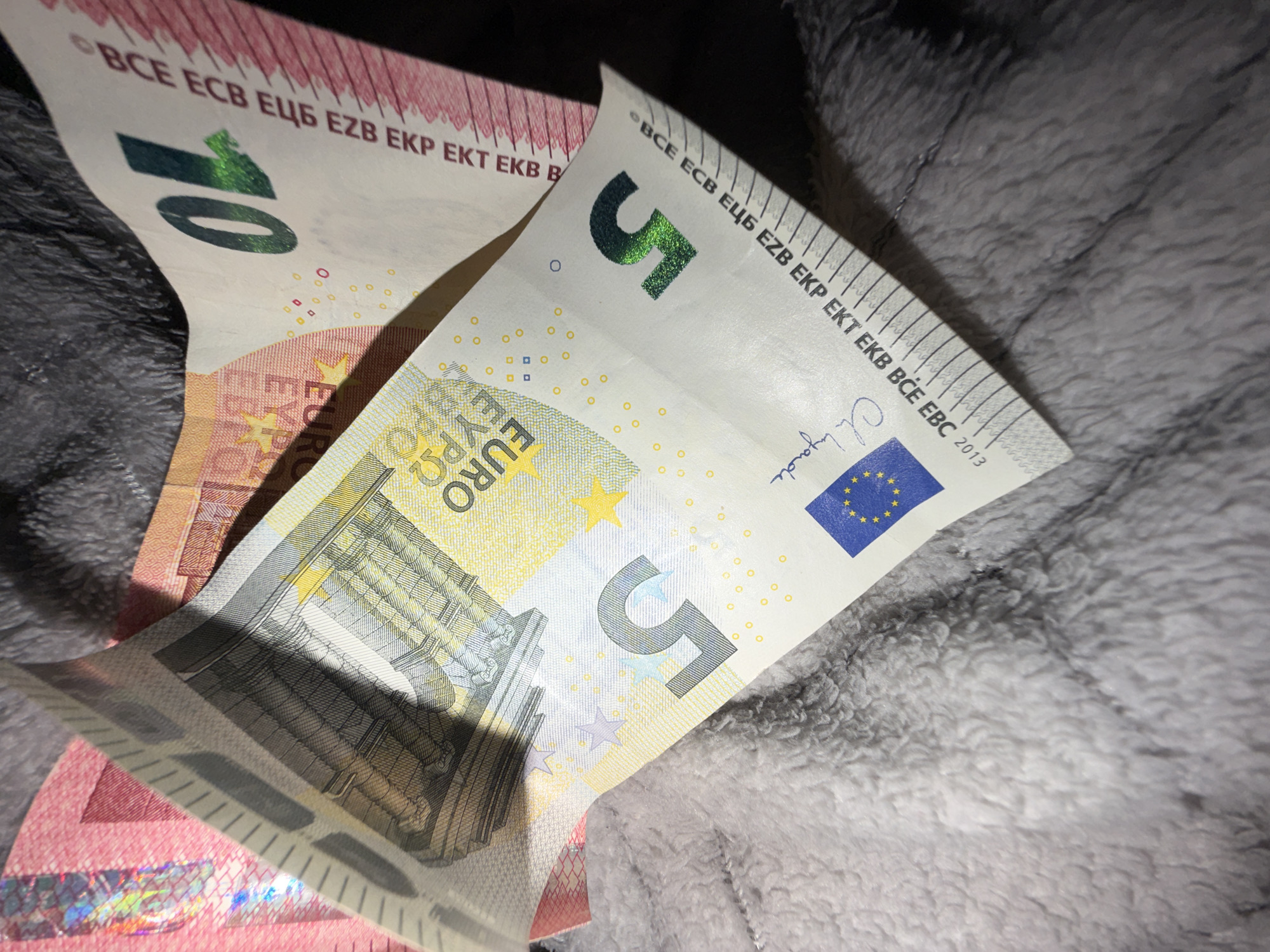 The image displays two euro banknotes, a 5 euro note and a 10 euro note, partially unfurled and resting on a textured gray surface, possibly a blanket or carpet. The banknotes are the primary subjects. The 5 euro note is more prominent, showing the number "5" in a metallic green ink and the European Union flag. The text "BCE ECB EЦБ EZB EKP EKT EKB BCE EBC 2013" is visible along the top edge of the note. The 10 euro note, partially obscured, shows the number "10" in a similar metallic green ink and the text "BCE ECB EЦБ EZB EKP EKT EKB B". The overall lighting suggests an indoor setting, possibly taken in the evening or with artificial light. The focus is on the details of the banknotes, highlighting their design elements and markings. There is no indication of people, specific activities, or a particular time of day or weather. The location context is Dorsten, Germany, but this is not visually discernible from the image itself. The scene is simple and focuses on the currency.