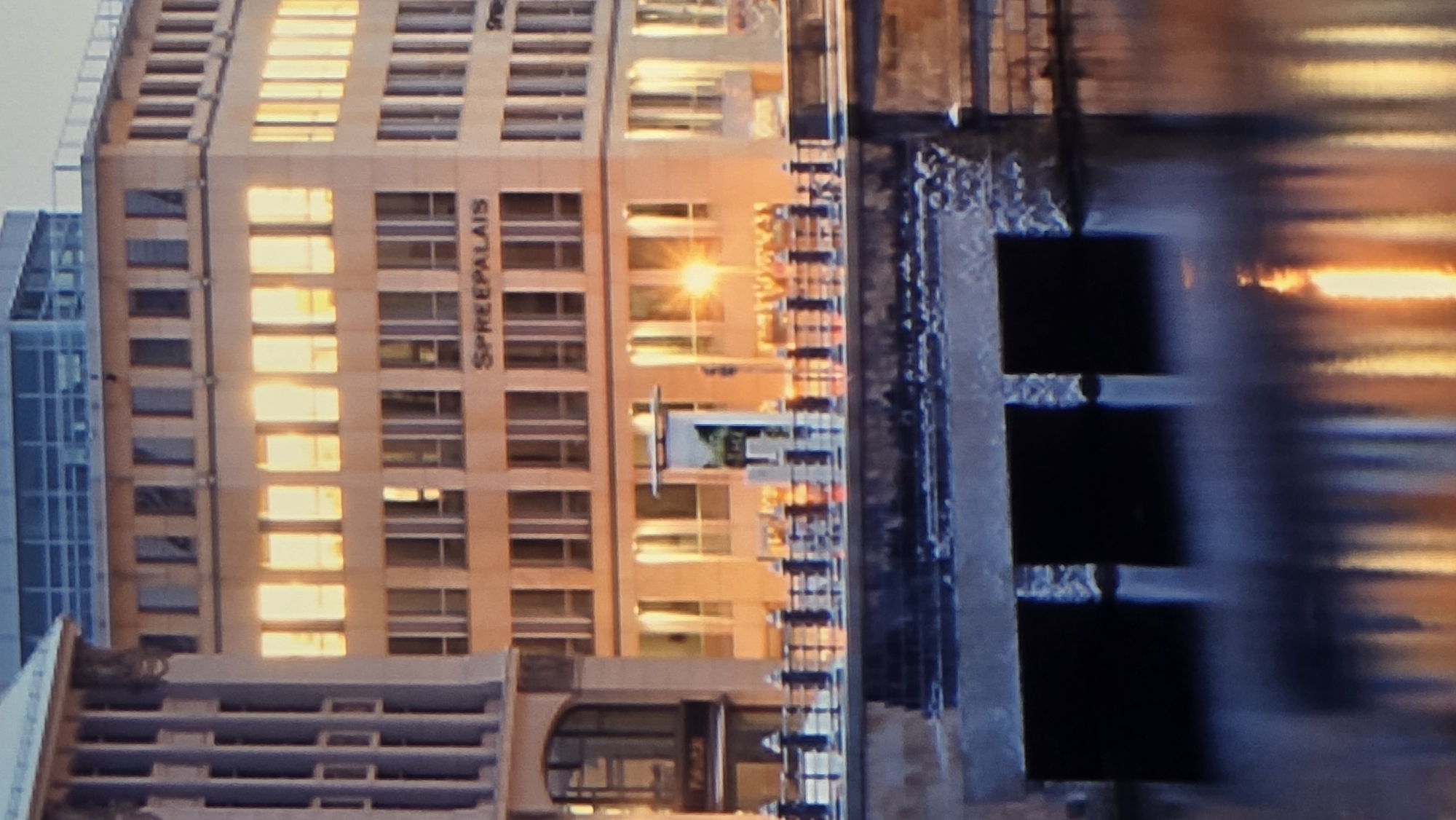 The image shows a city scene with a building on the right, featuring a sign that reads "SPREEPALAIS". The building has several floors with numerous windows, many of which are illuminated with a warm, yellow light, suggesting interior lighting or reflections. In front of the building, a bridge with ornate railings spans across a body of water. The water reflects the lights and structures of the scene, creating a shimmering effect. On the left side of the image, a portion of another building is visible, characterized by vertical columns. The overall atmosphere appears to be evening or dusk, given the lighting conditions. This image is not located in the city of Dobrich, Bulgaria.