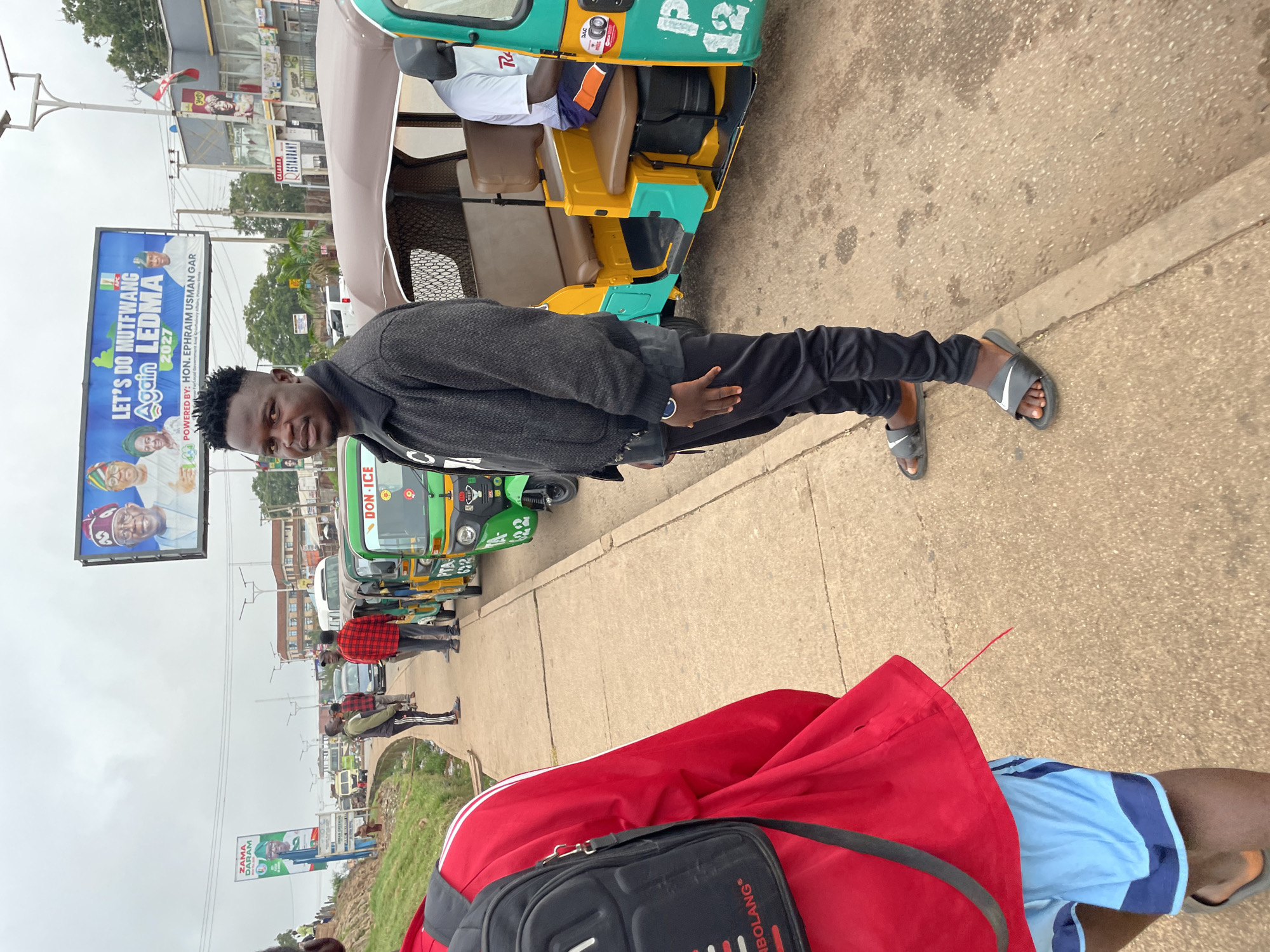 A young Black man stands on a sidewalk in Jos, Nigeria, looking at the camera. He is wearing a dark, textured jacket over a black t-shirt, black sweatpants, and grey Nike slides. To his right, a green and yellow auto-rickshaw is parked, with a man in a white t-shirt sitting in the passenger seat. Behind the auto-rickshaw, several other auto-rickshaws are visible, along with a white van and other vehicles. In the background, a large billboard displays images of political figures and campaign slogans. The sidewalk is made of concrete, and a drainage ditch runs alongside it. The sky is overcast.