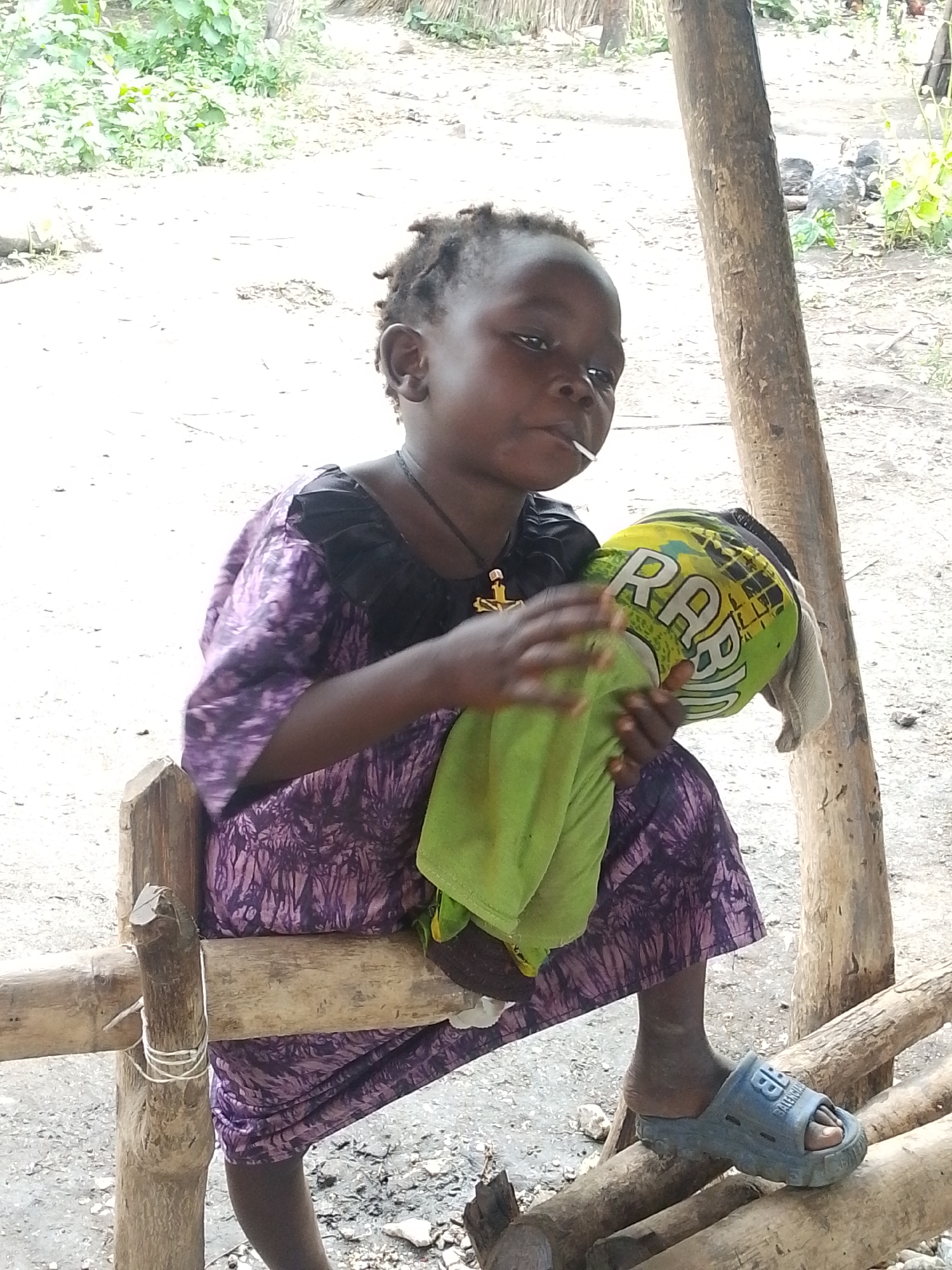 A young child, likely a girl, is seated on a rough wooden structure. She is wearing a purple patterned dress with black accents at the neckline and sleeves. Around her neck, she has a necklace with a cross pendant. Her right hand is holding a green cloth, and her left hand is cradling a yellow and green object with the letters "RA" visible on it. A white lollipop stick is in her mouth. On her right foot, she wears a blue sandal with a logo that appears to be "BALENCIAGA." The background suggests an outdoor setting with sandy ground, scattered rocks, and sparse vegetation. A thick wooden post is visible behind the child, to her right. The overall scene is consistent with a rural or semi-rural environment in Otulu, Nigeria.