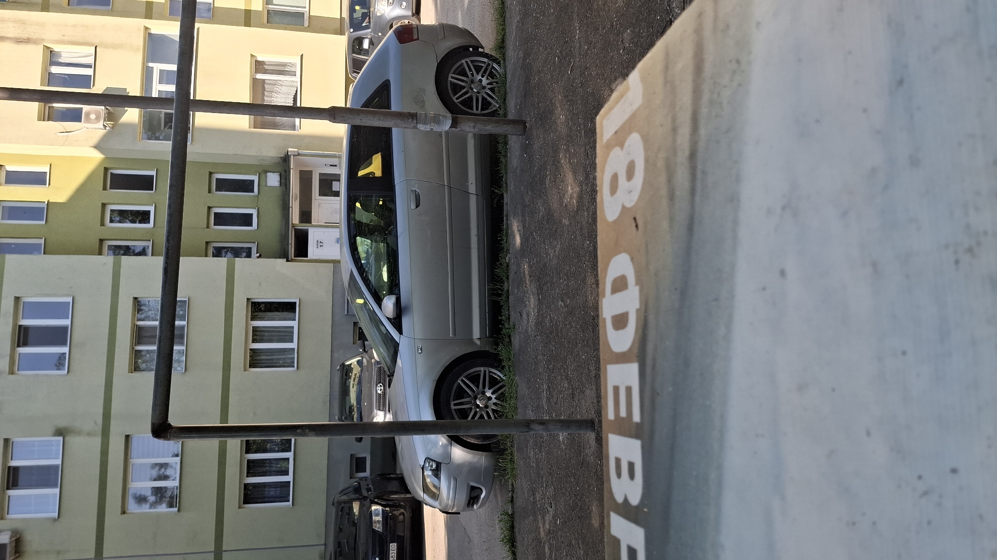 A silver Audi A3 hatchback is parked on a street in front of a light green apartment building. The car is facing to the right, and its driver's side is visible. The building has several windows and a light yellow accent. In the foreground, a blurred sign with white lettering is visible, with the partial text "февр 81" readable. Several other vehicles are parked in the background. The sun is shining, casting shadows on the scene.