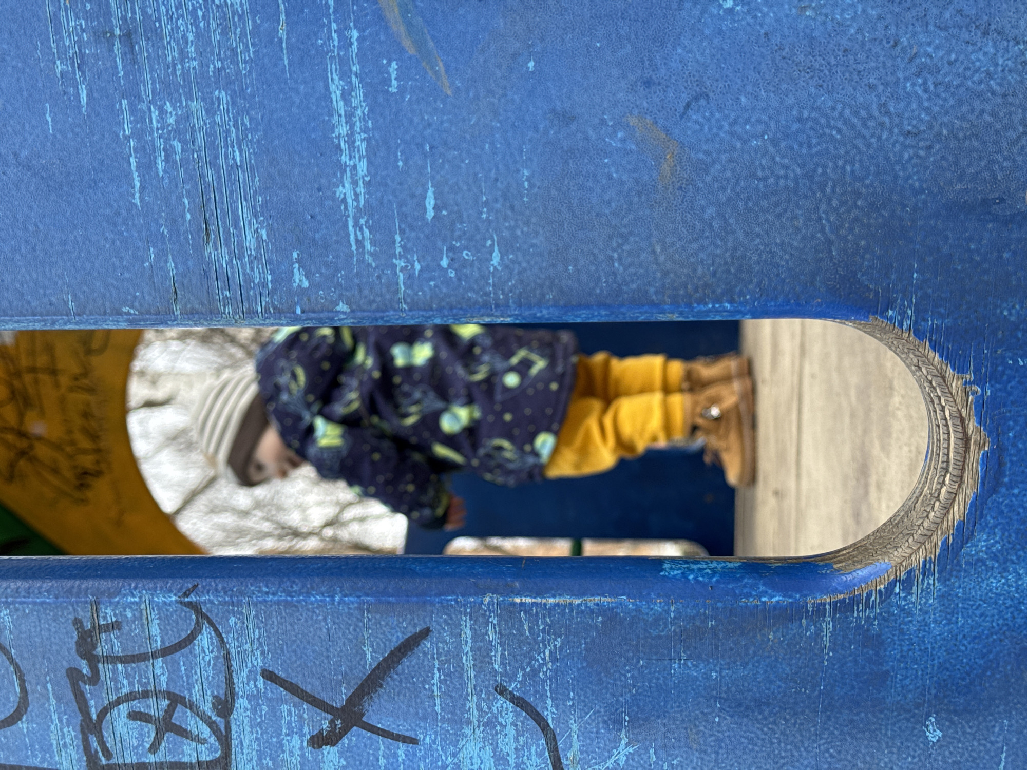 A close-up, vertically oriented photograph captures a child viewed through an oval-shaped opening in a weathered blue structure, likely playground equipment. The child, a toddler or young individual, is partially visible, wearing a dark blue jacket with a light pattern, yellow pants, and brown boots. A white and grey striped beanie covers their head. They appear to be standing or moving, with their head to the left and body extending right, looking downwards or away from the viewer.
The foreground and background are dominated by the blue structure, which exhibits significant wear and tear. Its surface is marked by numerous scratches, chipped paint revealing lighter blue tones or the underlying material, and a textured, slightly pebbled appearance in some areas. Dark, graffiti-like markings, including a prominent 'X' and other abstract strokes, are visible on the left side of the blue surface. The edges of the oval opening are also worn, showing layers and fraying of the material.
The scene appears to be outdoors during daylight hours, indicated by the bright, diffused light. The background seen through the opening is blurred, suggesting an outdoor setting, possibly a playground. The image has a slightly tilted perspective, emphasizing the texture and details of the blue structure and the glimpse of the child within.