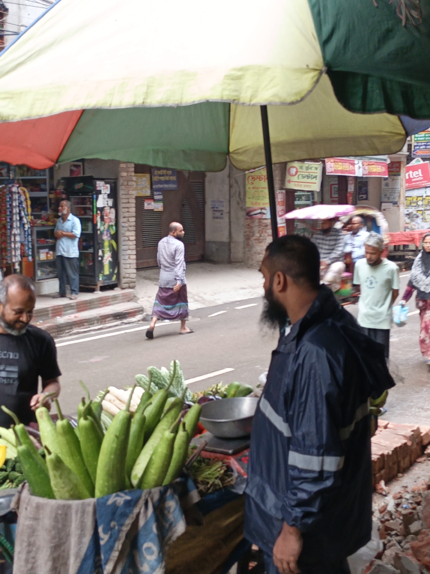 This photograph captures a vibrant street market scene, likely in an urban or town setting in South Asia, indicated by the visible Bengali script and local attire. The time of day appears to be daytime, under what seems to be an overcast sky, providing soft, diffused light.
In the foreground, a vendor stall is set up, prominently displaying fresh produce. A large quantity of long green gourds, possibly bottle gourds or ridge gourds, are arranged in a cloth-lined basket featuring blue and white patterns. Other vegetables like white radishes and green bitter gourds are also visible. Two men are present at the stall. On the left, a man with a beard, wearing a dark t-shirt with "ECHO" partially visible, is looking down at the produce. On the right, another bearded man wears a dark blue rain jacket with grey reflective strips, facing away from the camera and looking right, suggesting recent or impending rain. A metal bowl or scale is partially visible next to the vegetables. Providing shade over the stall is a large, colorful umbrella with sections of green, red, and light green/white fabric.
The midground reveals a street or narrow road with white line markings, where several people are in motion. On the left, inside a shop with shelves and clothing racks, a man in a light blue shirt stands. A bald man in a light shirt and a purple and blue patterned lungi walks across the street. Further to the right, a man in a light green t-shirt walks past, followed by a woman wearing a dark headscarf and a patterned dress. In the background, a rickshaw or cart with two men is also visible.
Buildings line the street, displaying various signs in Bengali script. One sign includes a tooth icon, indicating a dental clinic, and a red sign with "BARTEL" (likely Airtel) is also visible. The ground near the right edge of the frame shows scattered debris, including broken bricks.