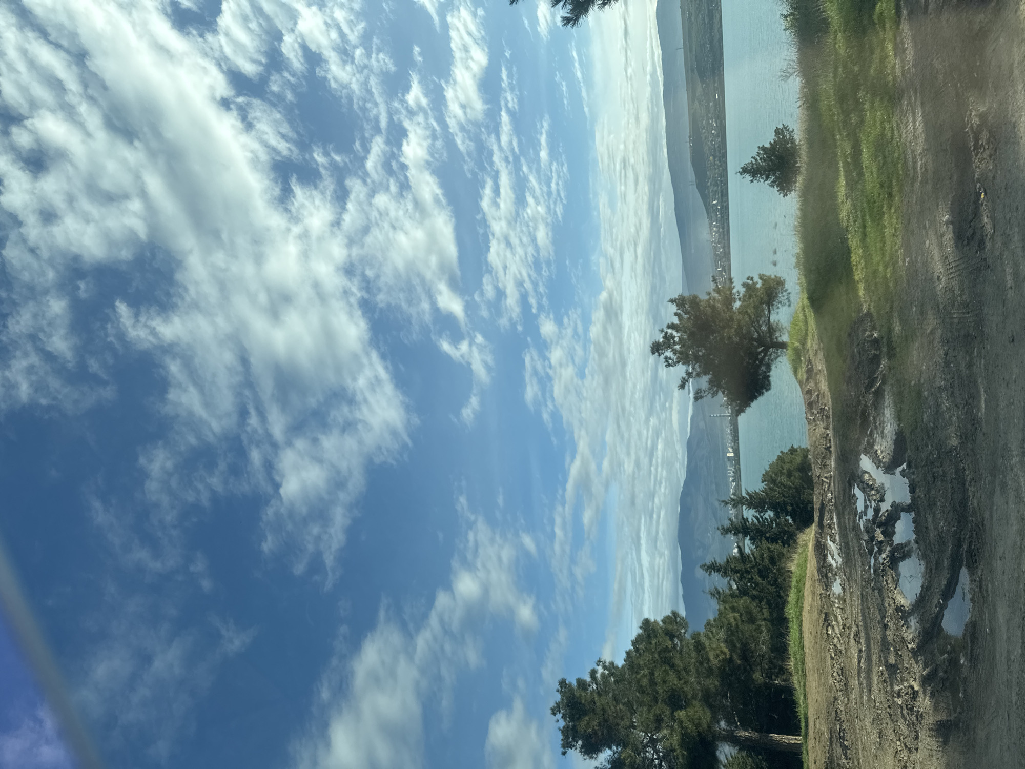 The image shows a landscape with a large body of water, possibly a lake or sea, visible in the distance. A shoreline with trees and vegetation separates the water from a dirt road in the foreground. The road appears muddy with puddles. The sky is blue with scattered white clouds. The image does not appear to be related to Tbilisi, Georgia.
