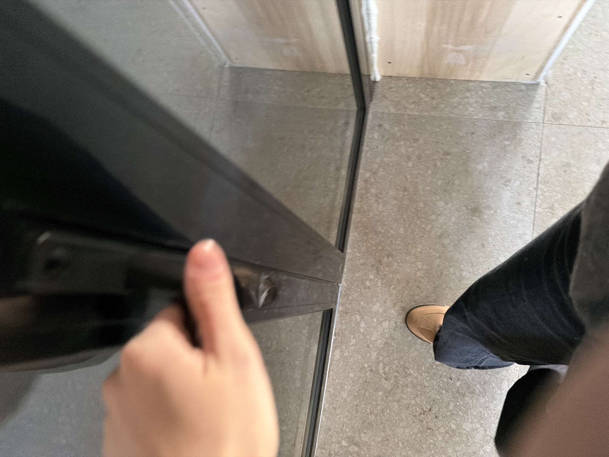 A first-person perspective image shows a hand opening a dark-framed door. The door is situated between two light-colored walls or panels, with a speckled gray tiled floor visible beneath. A person wearing dark denim jeans and a tan shoe is partially visible on the right side of the frame. The image suggests an indoor environment, but there is no specific information to determine its location in Chaikovsky, Russia.