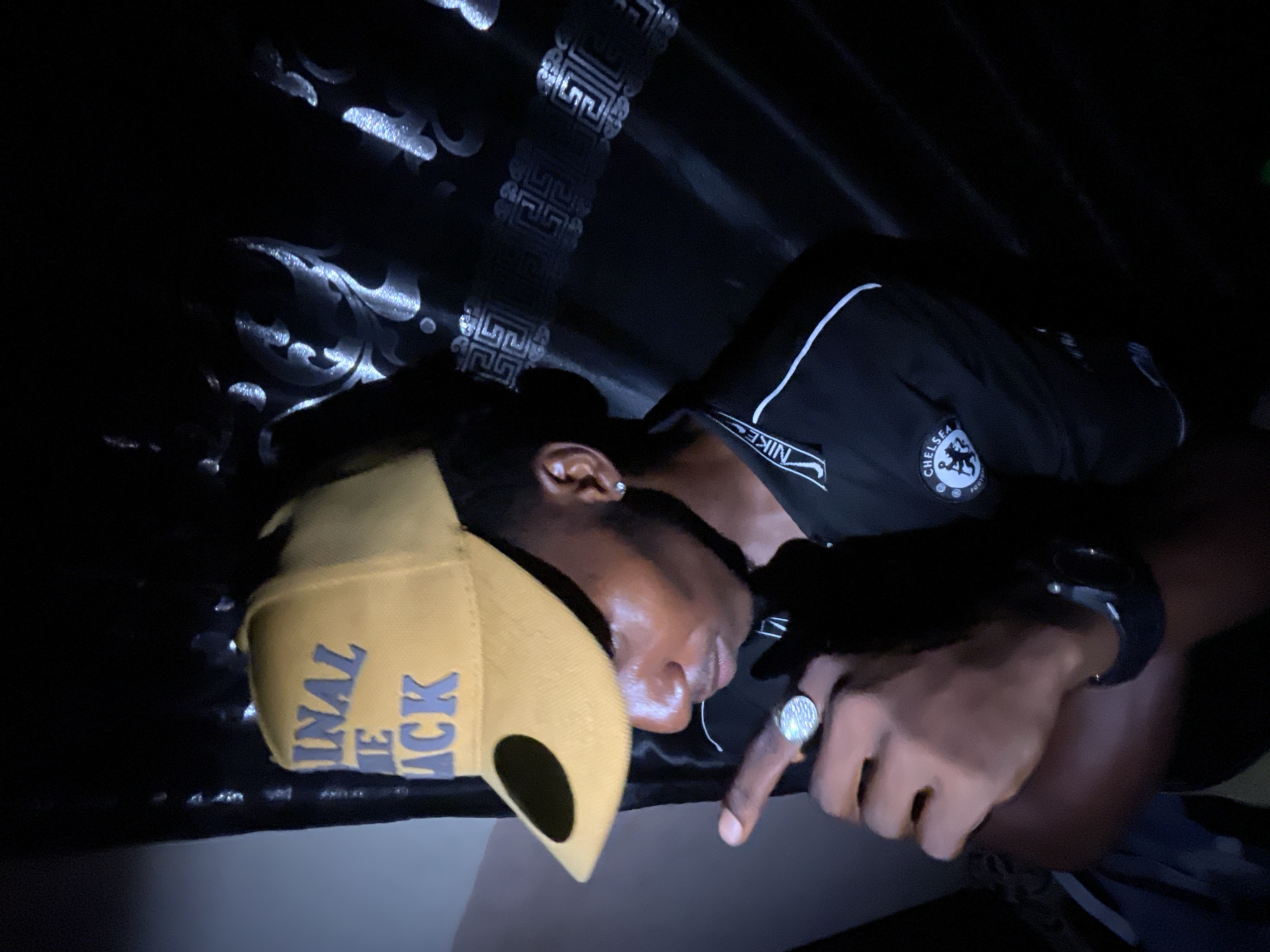 The image shows a person wearing a yellow cap with the words "Original Le Ack" and a black Chelsea FC shirt, lying down on a dark patterned surface. They are wearing a large ring on their index finger and a watch on their wrist. The environment appears to be dark and indoors. There is no information in the image to determine if this image was taken in the city of Lokoja, Nigeria.