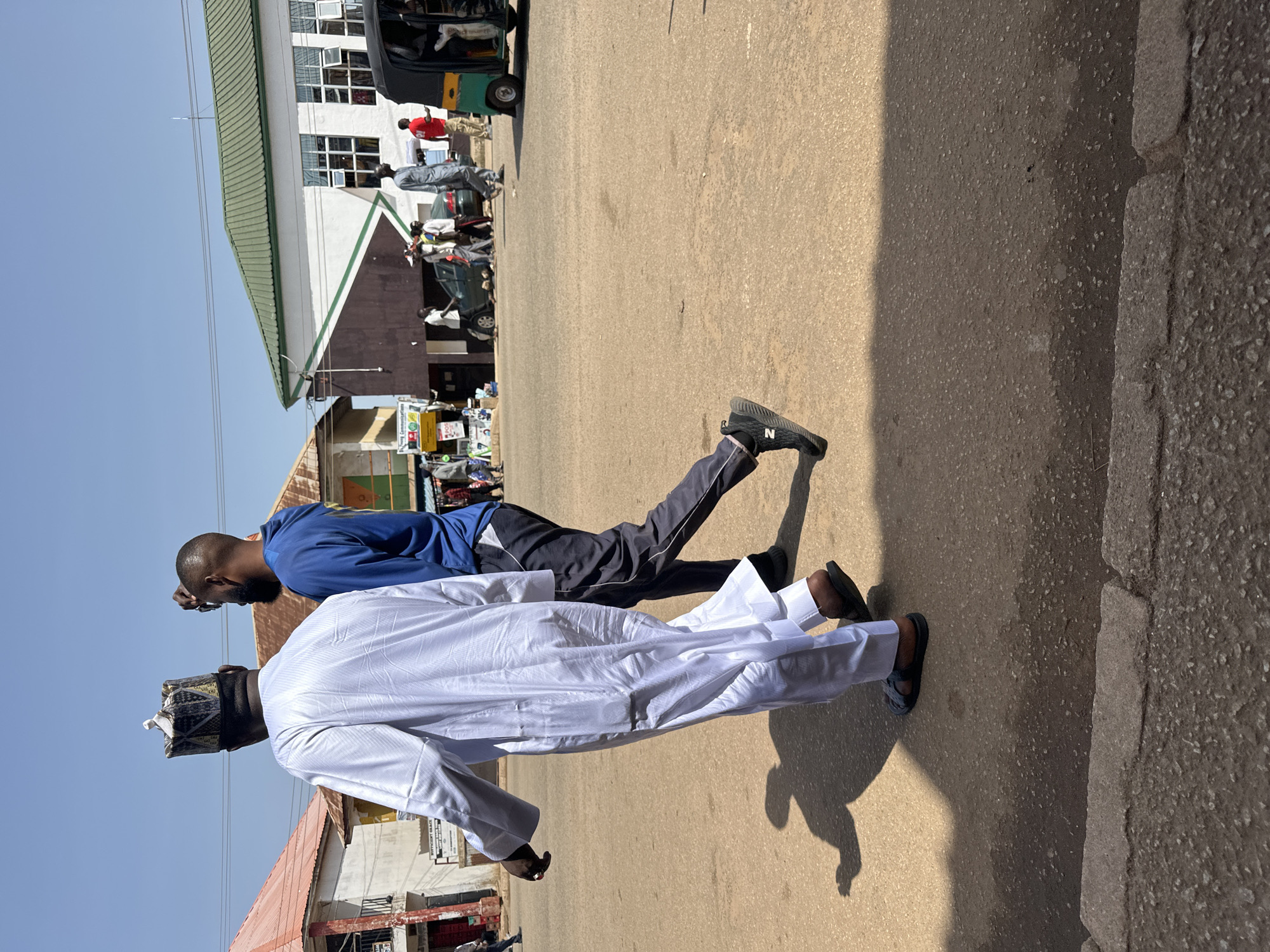 Two men are walking on a dusty street in Bukuru, Nigeria. The man on the left is wearing a white traditional robe and a patterned cap. The man on the right is wearing a blue hoodie and dark pants. They are walking away from the camera, casting long shadows on the ground. In the background, there are several buildings, including a white building with a green roof, and a colorful auto-rickshaw. Several other people and vehicles are visible in the distance, suggesting a busy urban environment. The sky is clear and blue.