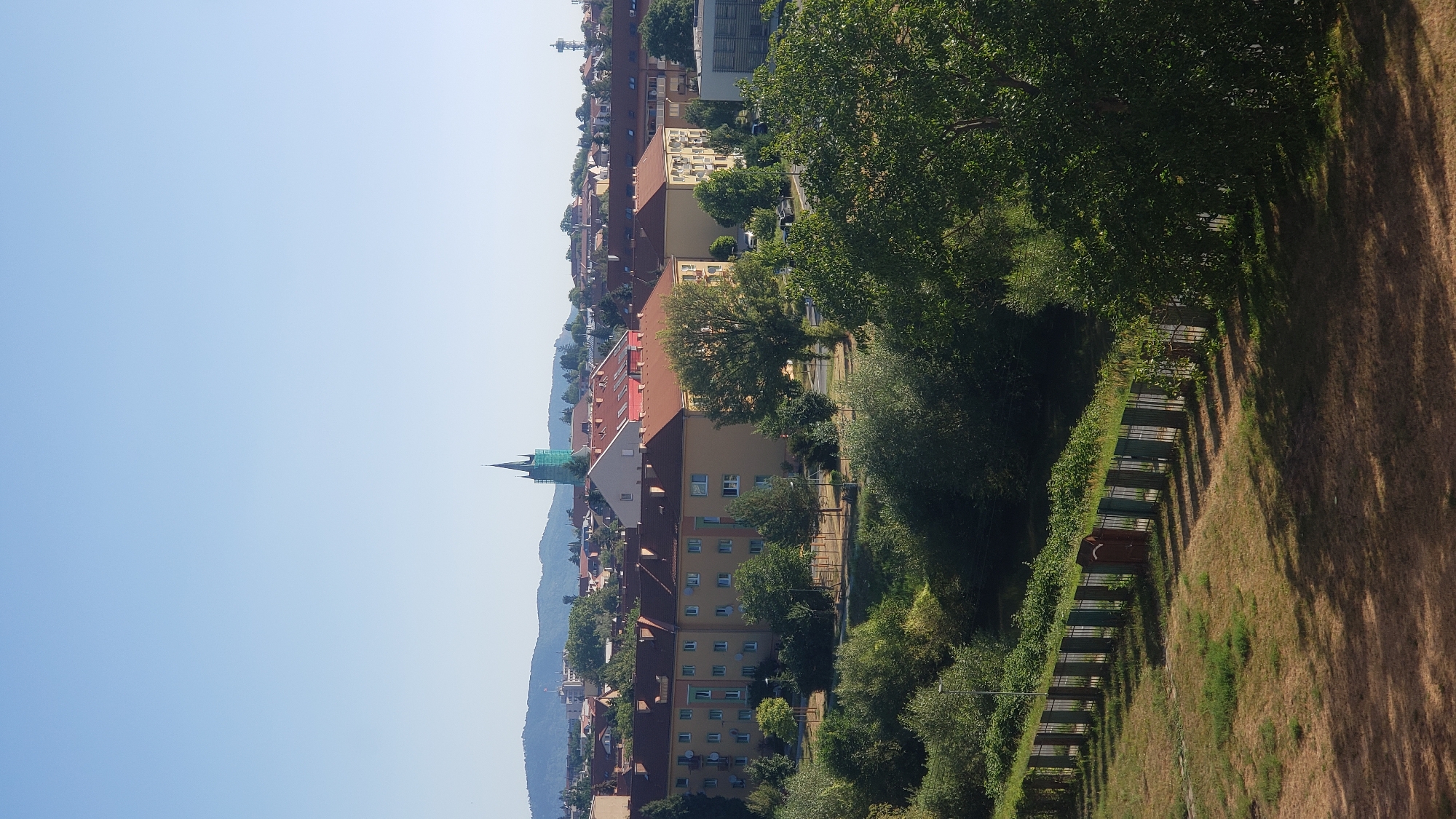 This is a vertical, medium-shot photograph taken from an elevated vantage point overlooking a cityscape under a clear, pale blue sky. In the distance, a range of hills is visible. The middle ground is dominated by a cluster of buildings with terracotta-tiled roofs, characteristic of European architecture. One prominent building is a yellowish-orange, multi-story apartment complex with visible windows and balconies. Further back, a slender, green-latticed spire, possibly part of a church or tower, rises above the rooftops. To the right, a water tower is visible.
The foreground of the image features a strip of dry, brown grass leading to a dark, metal fence with vertical bars. This fence runs horizontally across the lower portion of the frame, separating the grassy area from a more densely vegetated zone of trees and bushes. Shadows from the foliage are cast across the dry grass, indicating the direction of the sun. The overall scene suggests a residential area on the outskirts of a city, likely captured on a sunny day. Based on the architectural style, particularly the building with the terracotta roofs and the spire, this location is likely in the city of Prešov, Slovakia.