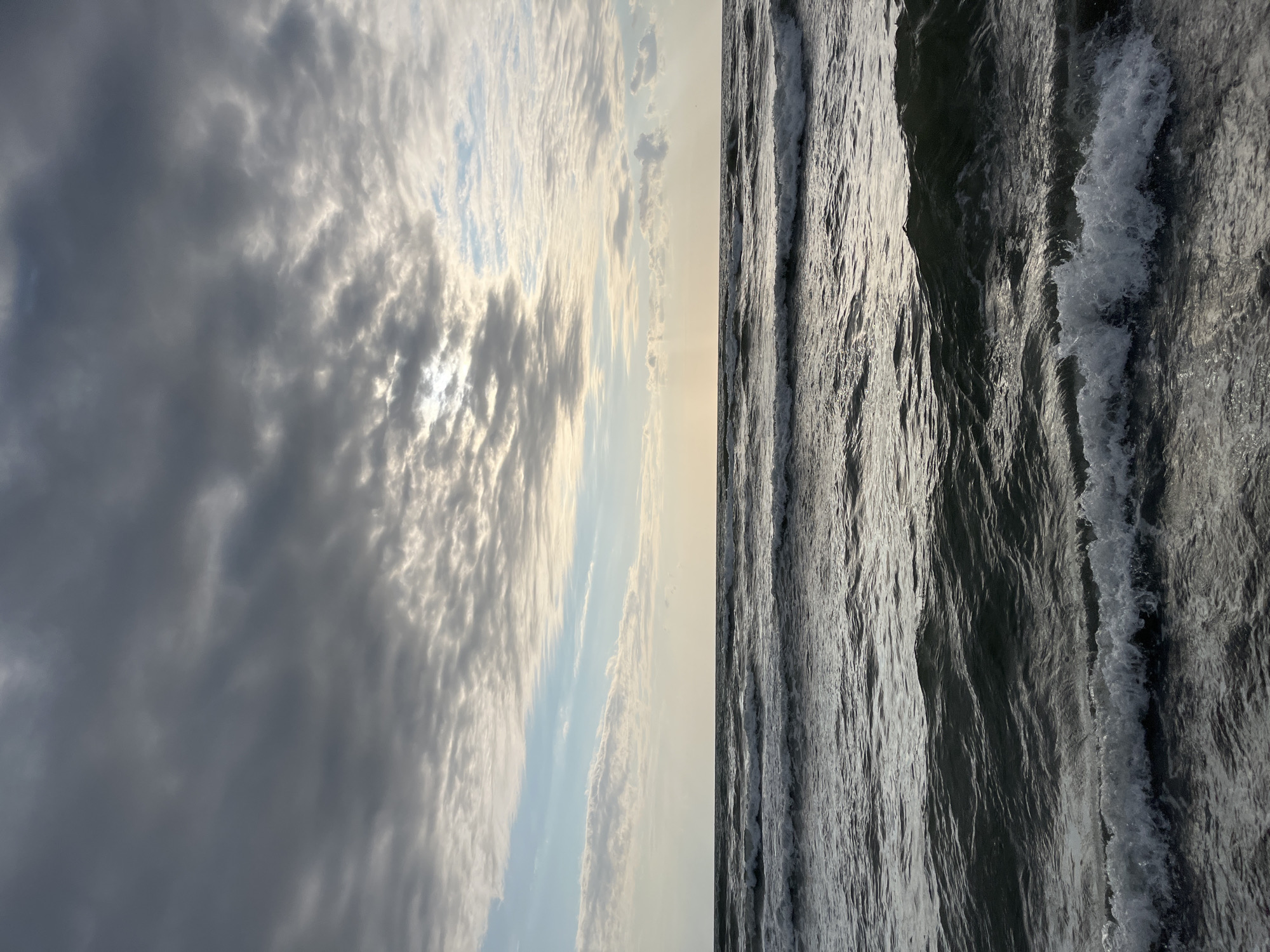 The image depicts a view of the Black Sea coast in Ureki, Georgia. The foreground is dominated by dark, choppy water with several small waves breaking, creating white foam along their crests. The water transitions to a calmer surface further out. The horizon line is a subtle strip between the sea and the sky. The sky is filled with a mix of dark, heavy clouds and lighter, more broken clouds. Patches of sunlight manage to break through the clouds, creating a slightly illuminated effect in the distance. There are no people or man-made objects visible in the image. The overall atmosphere is overcast and slightly dramatic, characteristic of coastal weather.