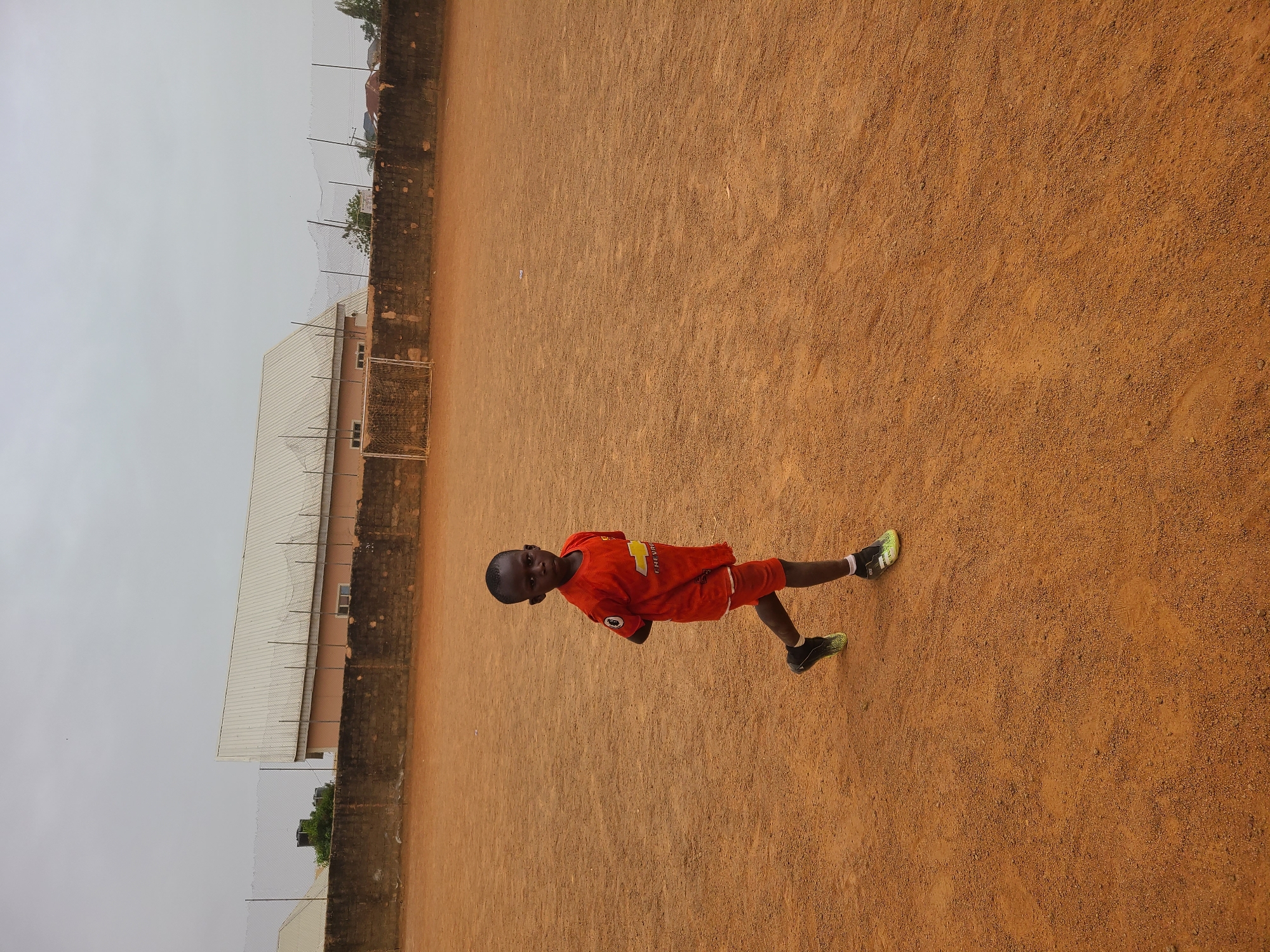 A young boy wearing a red soccer uniform and black and neon green cleats stands on a dirt field. He is facing the camera and looks to be in his early teens. Behind him is a building with a light-colored roof and a brick wall. The sky is overcast. The location is Barakin Lerawi, Nigeria.