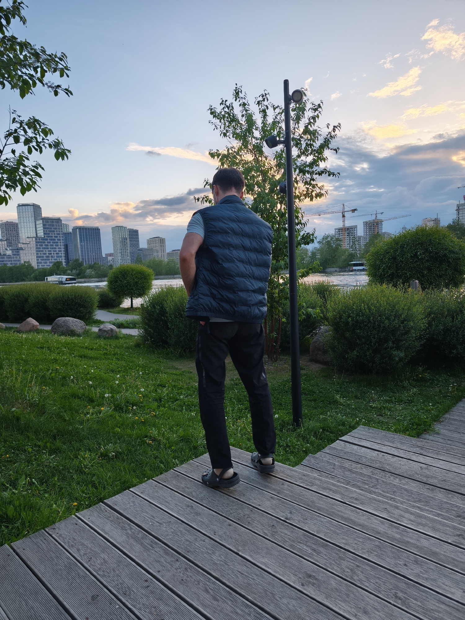 A man stands on a wooden deck in a park setting with a view of the Moscow skyline. He faces away from the camera, his hands in his pockets. He is wearing a dark blue vest over a light blue shirt, with dark trousers and sandals. The park has green grass, bushes, and a small tree. In the background are modern buildings, a river, and a partially cloudy sky with a hint of sunset. Construction cranes are visible in the distance. The location is in the city of Moscow, Russia.