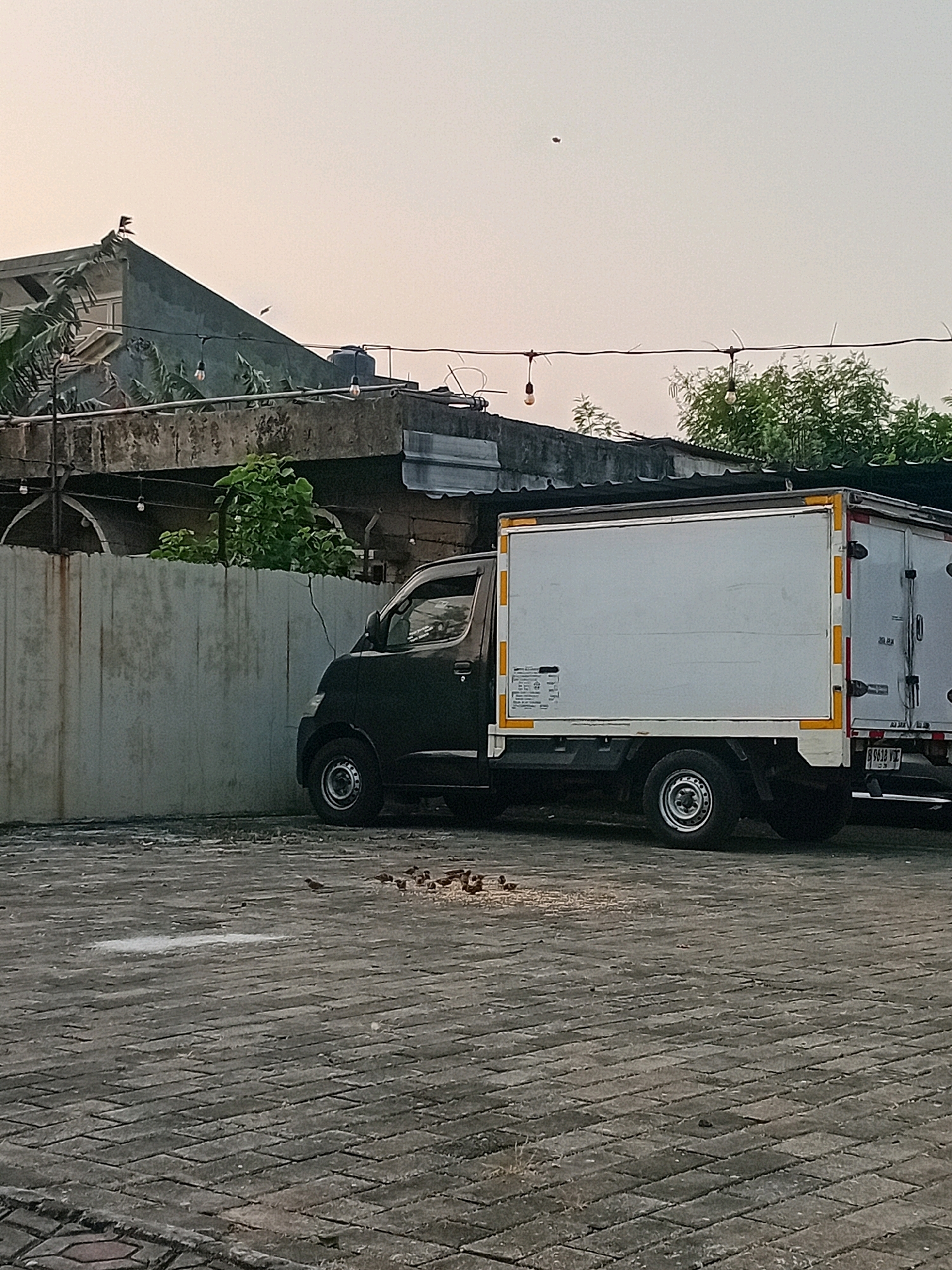 A black box truck is parked on a patterned brick surface in what appears to be an outdoor area, possibly a parking lot or loading dock, with a concrete wall to the left and buildings with roofs and foliage in the background. The truck has a white box body with yellow and orange accents on the rear and sides. Several small birds are gathered on the brick surface in front of the truck, pecking at scattered food particles. In the sky, a small, dark object, possibly a bird or insect, is visible. The overall lighting suggests it might be early morning or late afternoon. The presence of a water tank and exposed wiring with lights strung across the buildings indicates a functional, possibly commercial, setting within a city environment. Given the visual context, this scene is likely located in Tangerang, Indonesia, characterized by the common architectural styles and urban landscape.