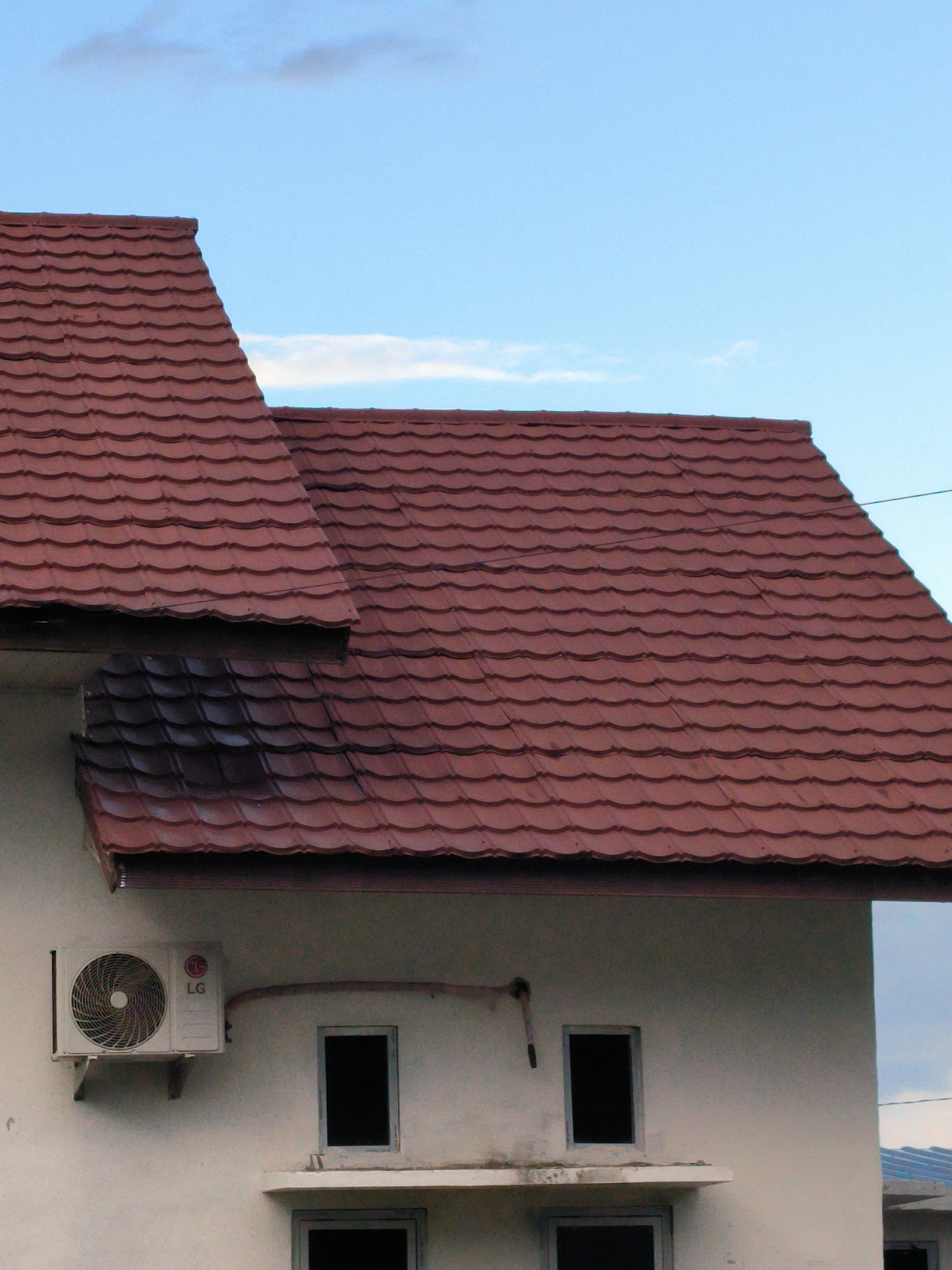 The image depicts the upper portion of a building in Lhokseumawe, Indonesia. The building has a distinctive reddish-brown tiled roof with a curved pattern. The exterior walls are painted in a light neutral color. An LG air conditioning unit is mounted on the wall. There are window openings present, but without glazing, appearing as dark rectangular holes. The sky above is blue with sparse clouds.