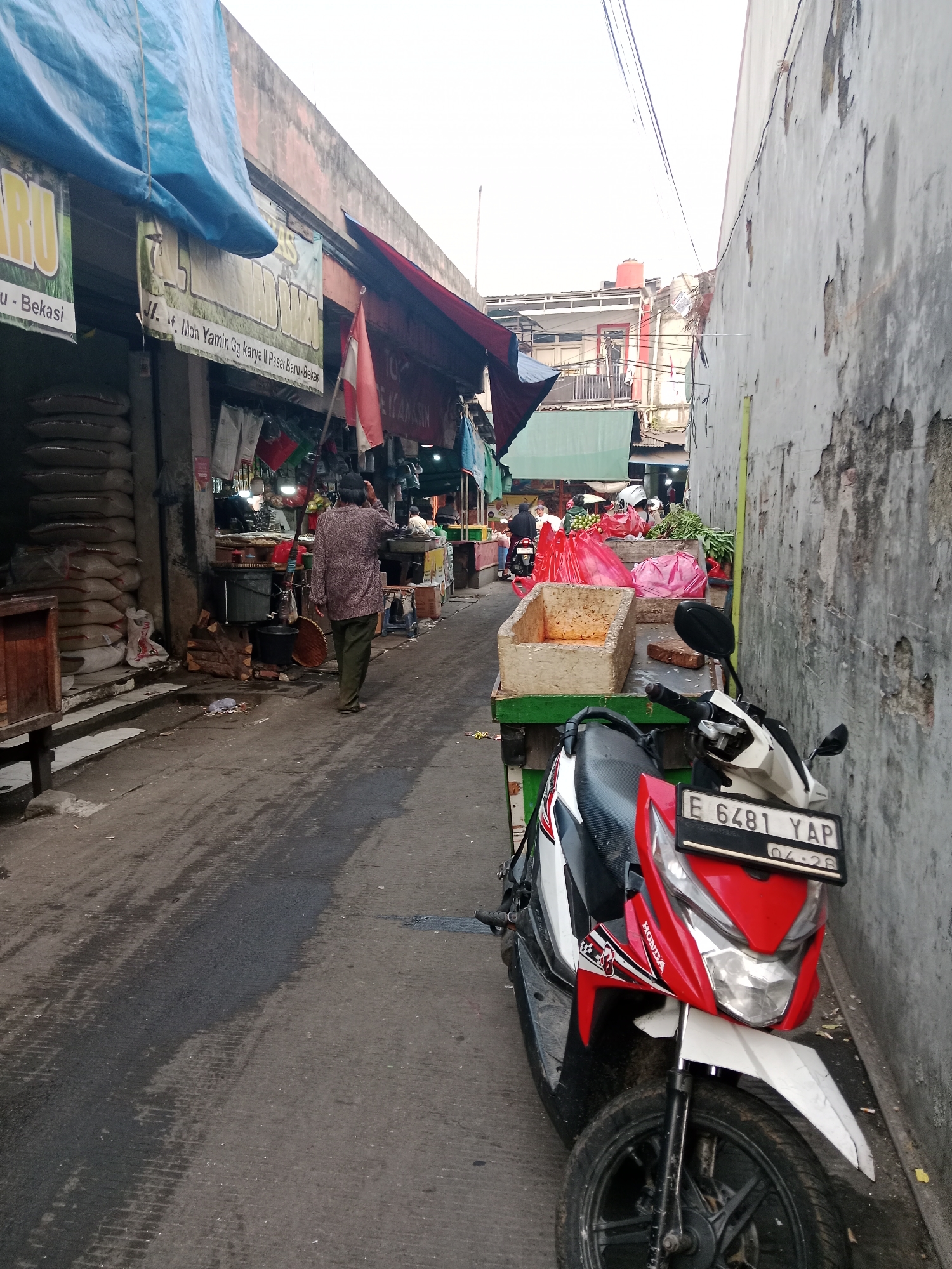 The image depicts a narrow street market in Bekasi, Indonesia, with stalls lining both sides. In the foreground, a red and white Honda scooter is parked, its license plate reading "E 6481 YAP." The street is wet and shows tire tracks, indicating recent rain or cleaning. On the left, market stalls display large sacks of what appears to be rice or grains, covered by blue tarps. A person in a patterned shirt and green pants walks away from the camera down the street, towards the bustling market activities.
Further into the market, more stalls are visible, with various goods, including produce like red tomatoes and green leafy vegetables, displayed in baskets and plastic bags. Some stalls have awnings and banners with text that is difficult to read. Red and white flags are also visible, adding to the market atmosphere. The buildings in the background appear to be a mix of residential and commercial structures, some with multiple stories and exposed electrical wiring. The overall environment suggests a vibrant, active marketplace. The wall on the right side of the image is weathered and shows signs of age and disrepair.