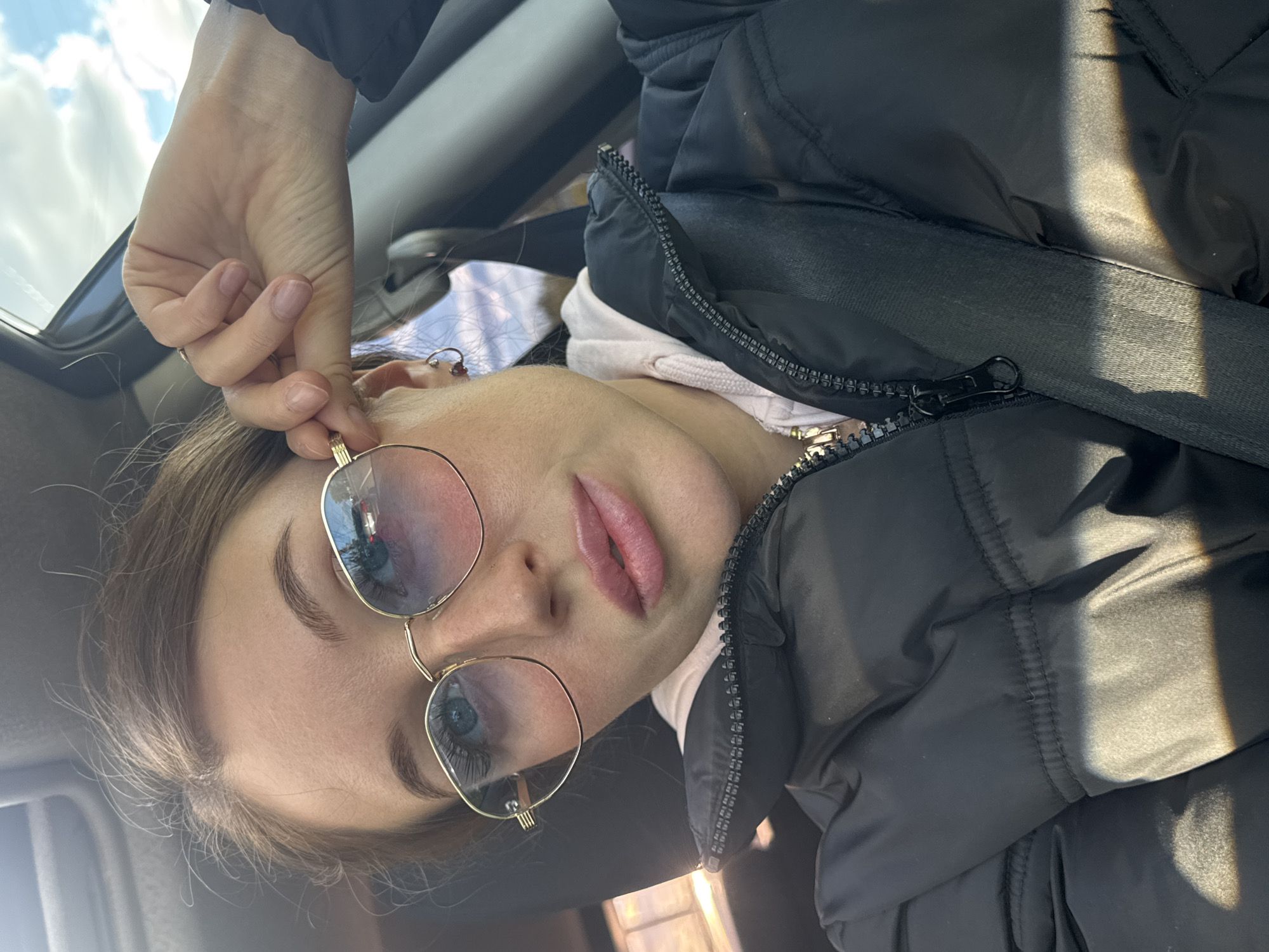 A young woman wearing gold-rimmed glasses, a pink hooded sweatshirt, and a black puffer jacket poses for a selfie in a vehicle. Her left hand is near her face, adjusting her glasses. The background includes a car interior and a glimpse of the sky. Location details (Chaikovsky, Russia) cannot be determined from visual evidence alone.