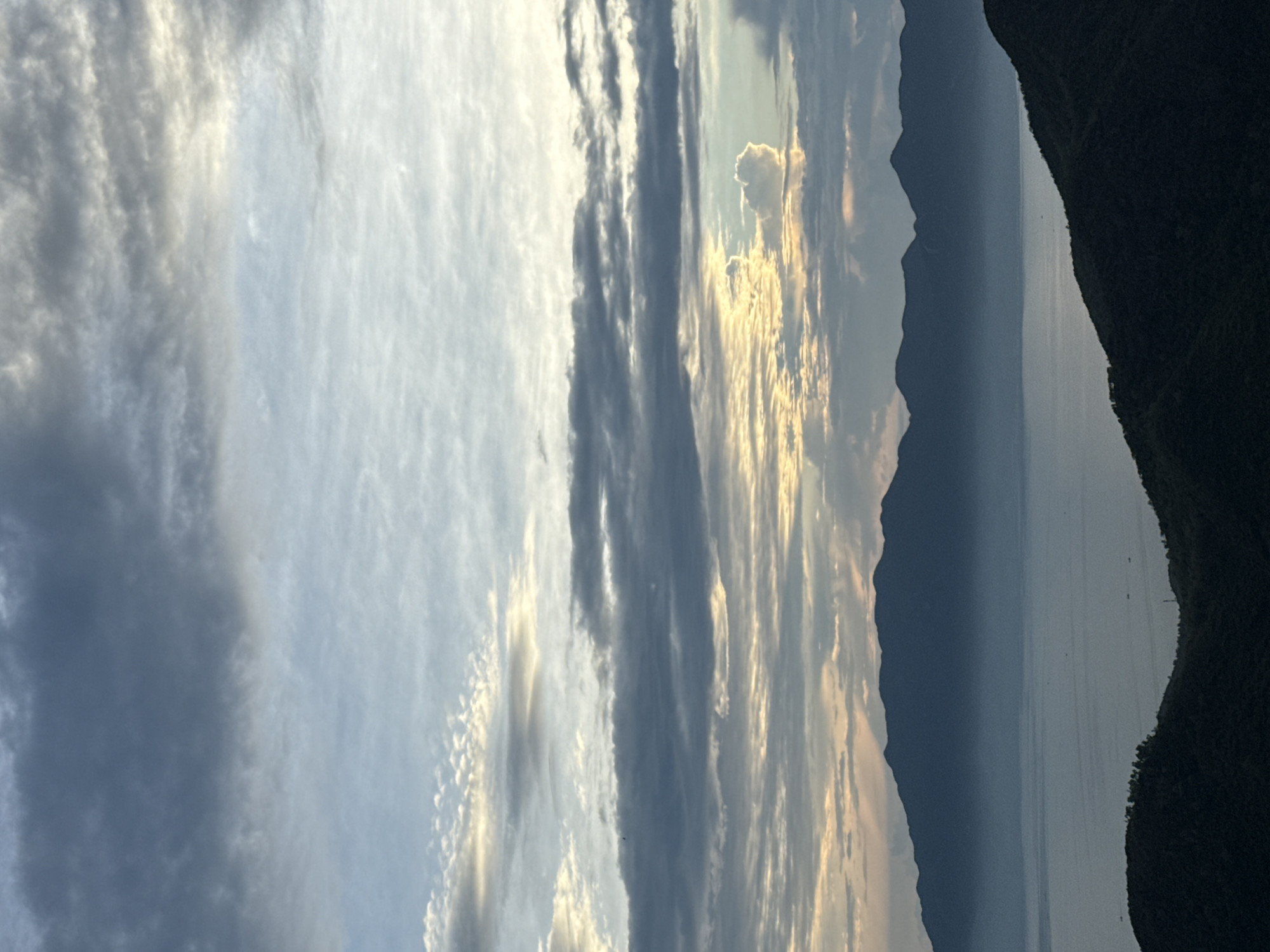 The image is a landscape featuring a sky with a variety of clouds ranging from light and fluffy to darker, more defined formations. Sunlight filters through some of the clouds. A mountain range is silhouetted against the lighter sky, with a body of water appearing to separate the viewer from the mountains. A darker land mass, possibly a hillside or more mountains, is visible to the right side of the image. The location is near Nha Trang ward, Vietnam. There are some electric pole outlines over the body of water.
