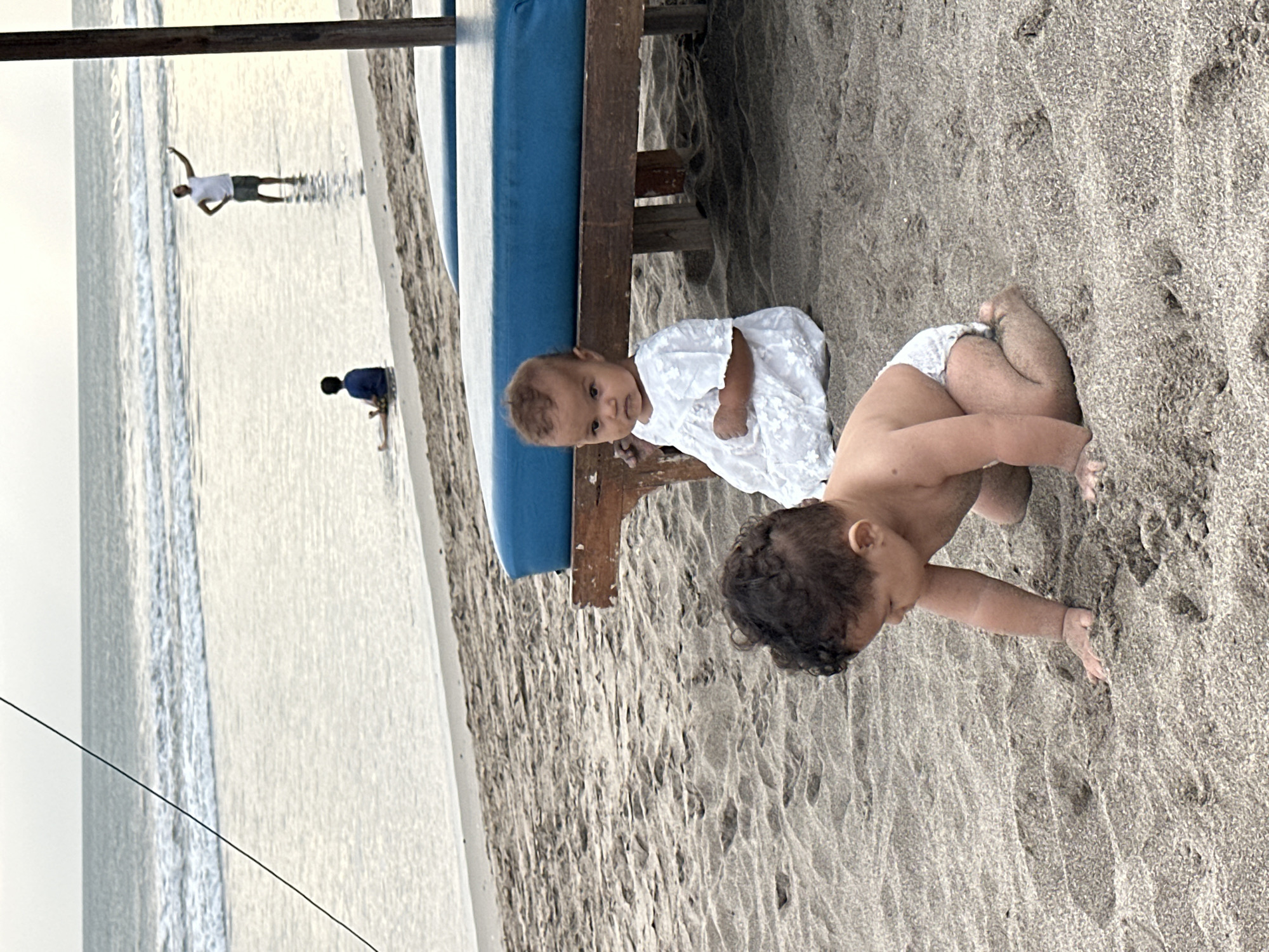 An eye-level, medium shot captures two babies on a sandy beach in North Kuta, Indonesia, during overcast daylight. In the foreground, a baby with dark, curly hair is on its hands and knees, exploring the sand. To its right, a baby wearing a white dress sits on the sand, looking towards the camera with a neutral expression. Behind the babies, a wooden beach lounger with a blue cushion is visible. In the background, the ocean meets the shore, with gentle waves rolling in. Further in the distance, two figures are visible in the shallow water: one person sits on the beach, while another stands and gestures with their right arm raised. The overall atmosphere is calm and serene, with the muted light contributing to a peaceful beach scene.
