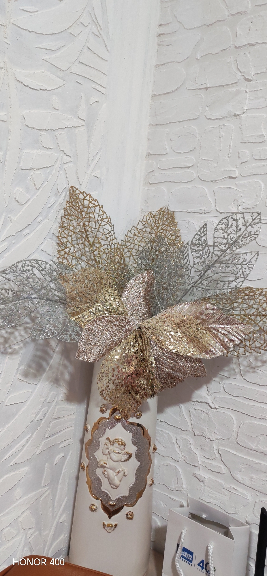 A white vase with a decorative angel and golden accents is displayed against a textured white wall, adorned with gold and silver artificial floral arrangements. Part of a white gift bag with blue branding is visible at the bottom right. The image was taken in Noviy Urengoy, Russia, according to the text on the bag.