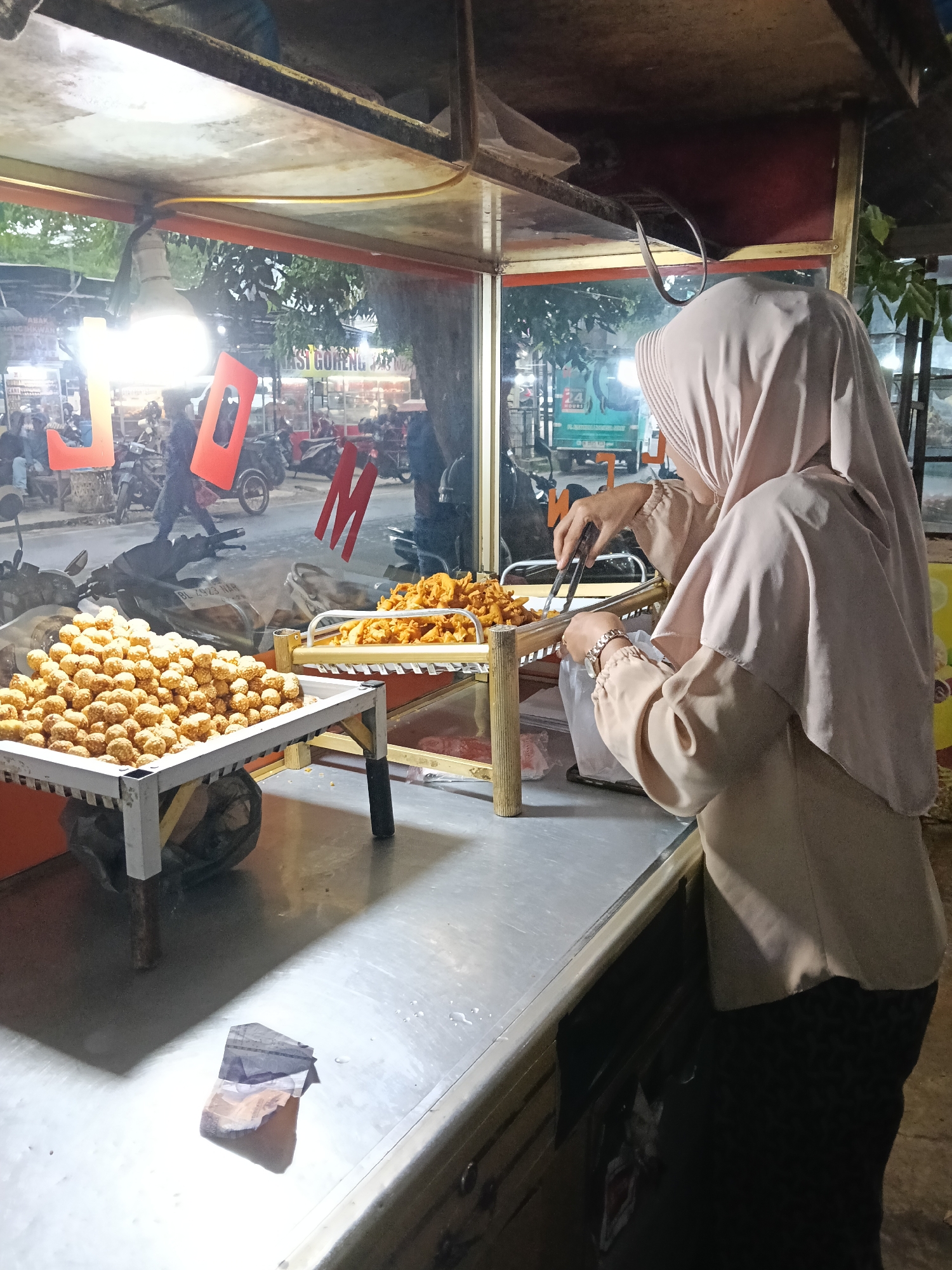 The image shows a street food vendor in Lhokseumawe, Indonesia. A person wearing a hijab and light-colored clothing is working at a food stall. The stall has a glass display case containing fried snacks. On the left is a tray piled high with round, golden-brown treats, and to the right, a tray holds smaller, fried items. The vendor is using tongs to handle the food. In the background, visible through the glass, is a street scene with motorbikes, other people, and signage. A piece of Indonesian currency is visible on the counter. The scene is lit by artificial lights.