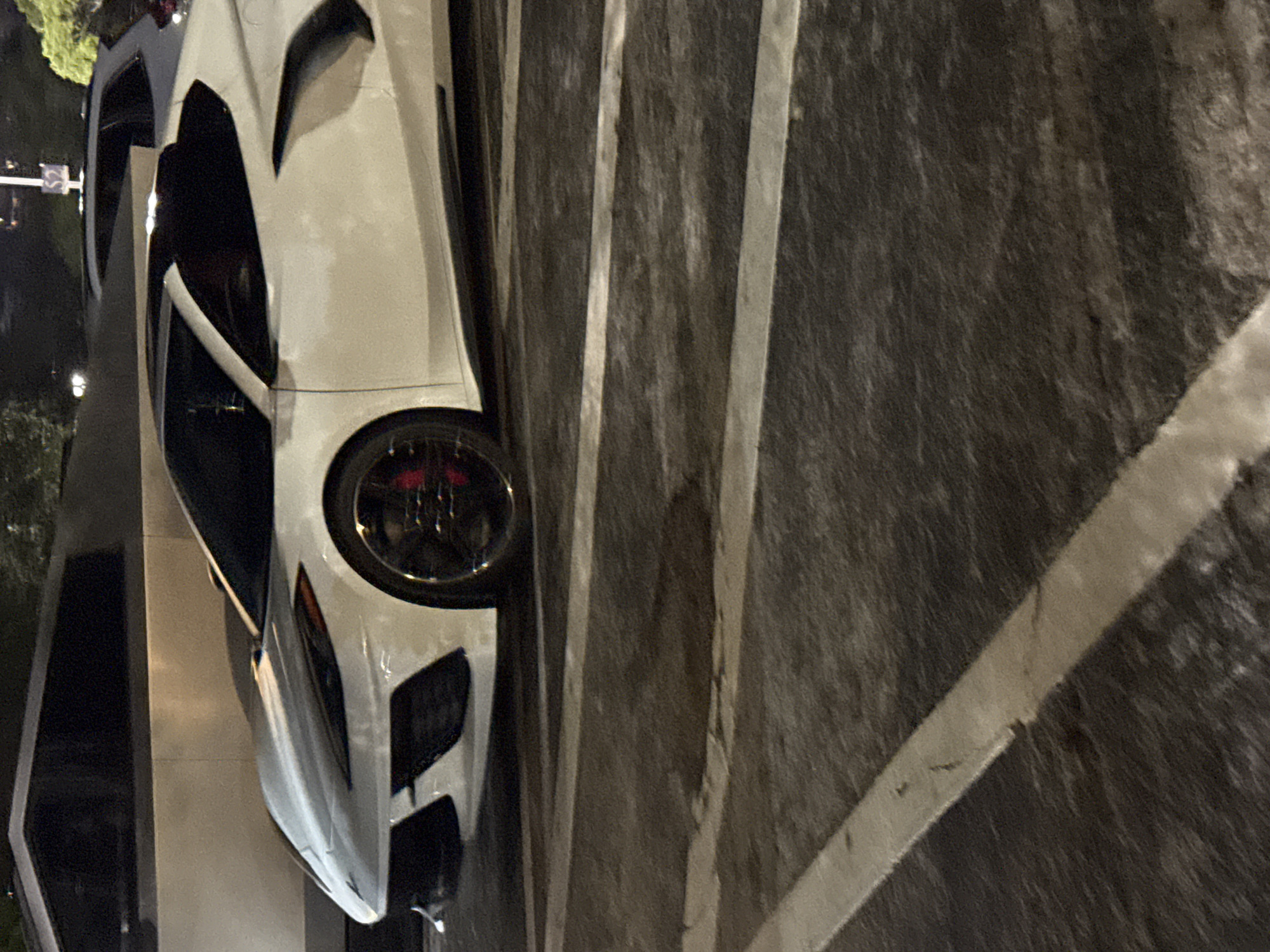 The image displays a white high-performance sports car, likely a Lamborghini, parked in an outdoor lot at night. The car is positioned at an angle, showcasing its sleek, aggressive front design with sharp lines and prominent air intakes. One of its front wheels is visible, revealing a dark rim and striking red brake calipers.
The setting appears to be a parking space, indicated by the distinct white lines painted on a dark asphalt surface. The lighting is dim, suggesting it's nighttime or late evening, with some distant artificial lights faintly visible in the background on the left side of the frame. The background also includes dark structures or parts of other vehicles, and what might be foliage further back. No people are visible, and the car is stationary.
Notable details include the car's pristine white paint contrasting with the dark background, and its futuristic, angular bodywork. The texture of the asphalt pavement is rough, and the white parking lines show signs of wear. In the far distance on the left, a blurry white sign or street light post is barely discernible, but any text on it is unreadable due to distance and blur. The overall impression is one of a powerful, luxury vehicle resting in an urban or suburban nocturnal environment.