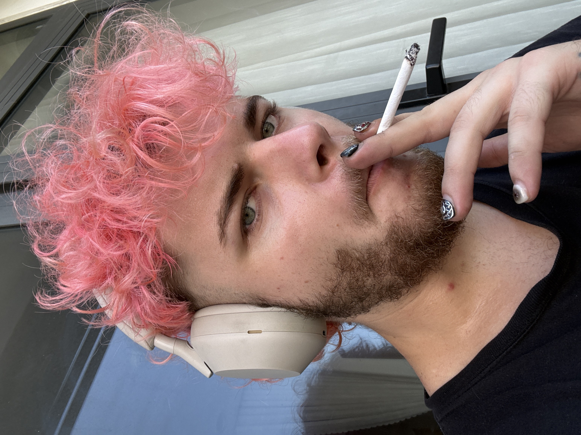 A young man with pink curly hair is holding a lit cigarette to his mouth. He has a light beard and light eyes. He is wearing headphones around his neck and his fingernails are painted with a design. He is wearing a black t-shirt. The background is a grey door and white blinds. There is no indication of the location being Đà Nẵng, Vietnam.