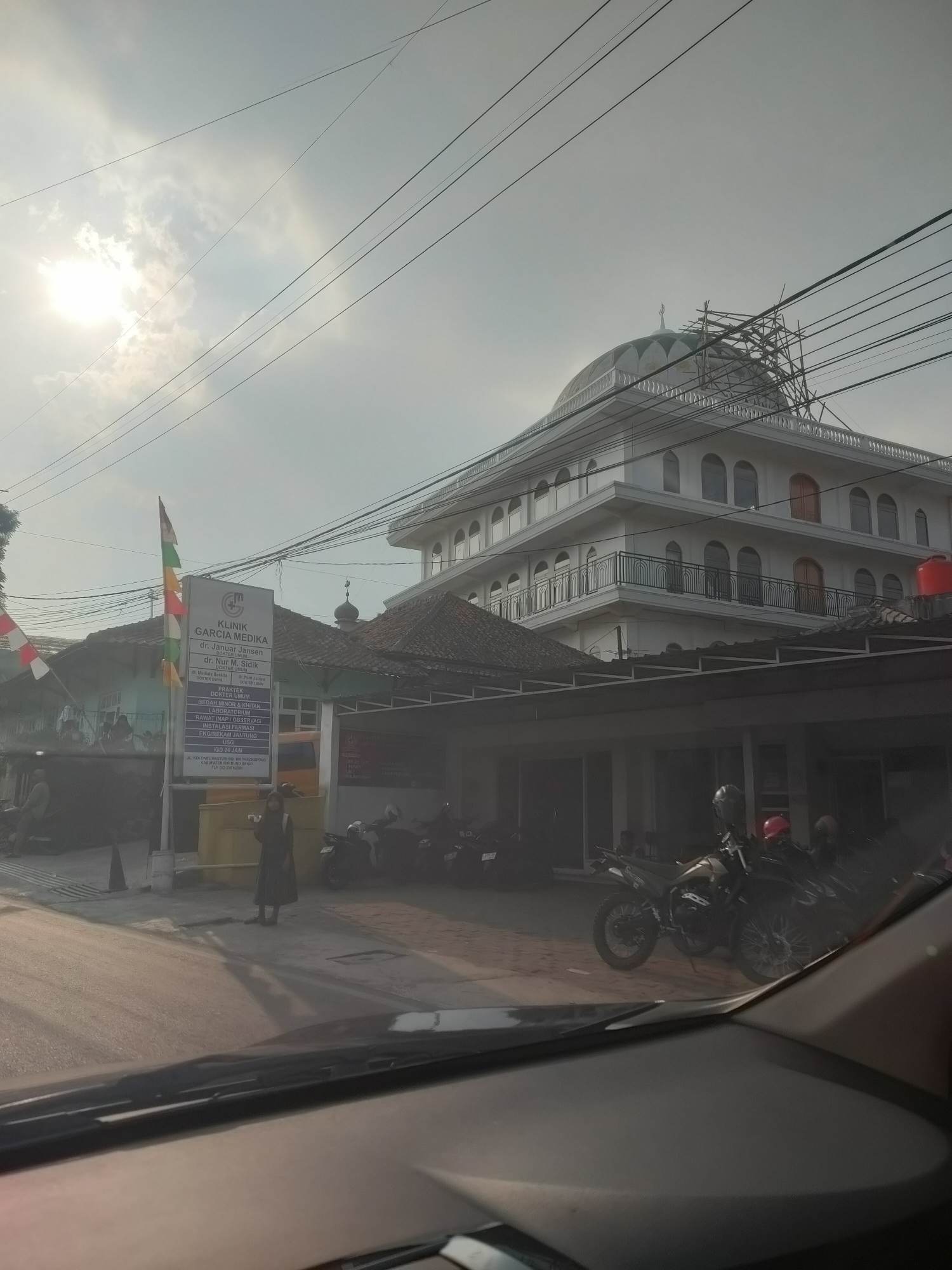 The image captures a street scene in Parongpong, Indonesia, with a prominent white building featuring a large dome under construction, suggesting it may be a mosque or a similar religious structure. In the foreground, a clinic sign for "KLINIK GARCIA MEDIKA" is visible, listing doctors' names and services, including "PRAKTEK DOKTER UMUM," "LABORATORIUM," and "IGD 24 JAM." Several motorcycles are parked on the street, and a person is standing near the clinic sign. The sky is partly cloudy with bright sunlight. The view appears to be taken from inside a vehicle, as evidenced by the windshield in the bottom of the frame. The overall impression is a functional, everyday scene in a built-up area of the city.