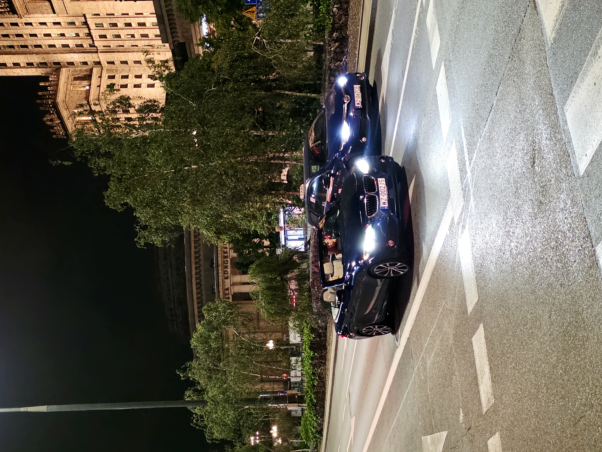 An exterior, eye-level, low-angle shot depicts a dark blue convertible BMW driving on a city street at night. The car's headlights are on. A person is visible in the driver's seat. In the background, a large, ornate, light-colored building with many windows and decorative architectural elements looms, identified by signage as the Palace of Culture and Science. Trees with green foliage line the street between the road and the building. The street is paved and marked with white lines and pedestrian crossings. Another dark blue car, a hatchback, is driving in the lane to the right of the BMW. The hatchback has a license plate that reads "WZ 067CE." The BMW's license plate reads "WZ 002JS." The scene appears to be Warsaw, Poland, near the Palace of Culture and Science.