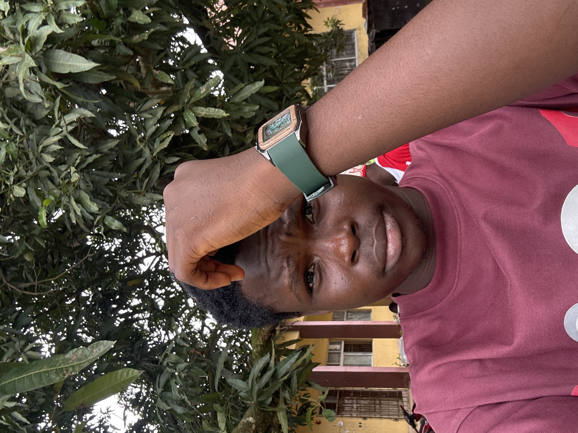 A close-up, high-angle shot captures a young Black person from the chest up, posing for a selfie. Their right hand is raised and touching their forehead, drawing attention to their facial expression of mild distress or concern. They are wearing a maroon t-shirt. A green-strapped watch with a gold-toned bezel and a green face is visible on their right wrist.
The background consists of lush green foliage from a tree, with a yellow building visible behind it. The building appears to have windows and doors, suggesting an urban or semi-urban setting. The overall lighting suggests an outdoor environment, possibly during the daytime. The image is sharp and in focus, highlighting the subject's features and the details of their attire and accessories. The location is identified as Ihiagwa, Nigeria.