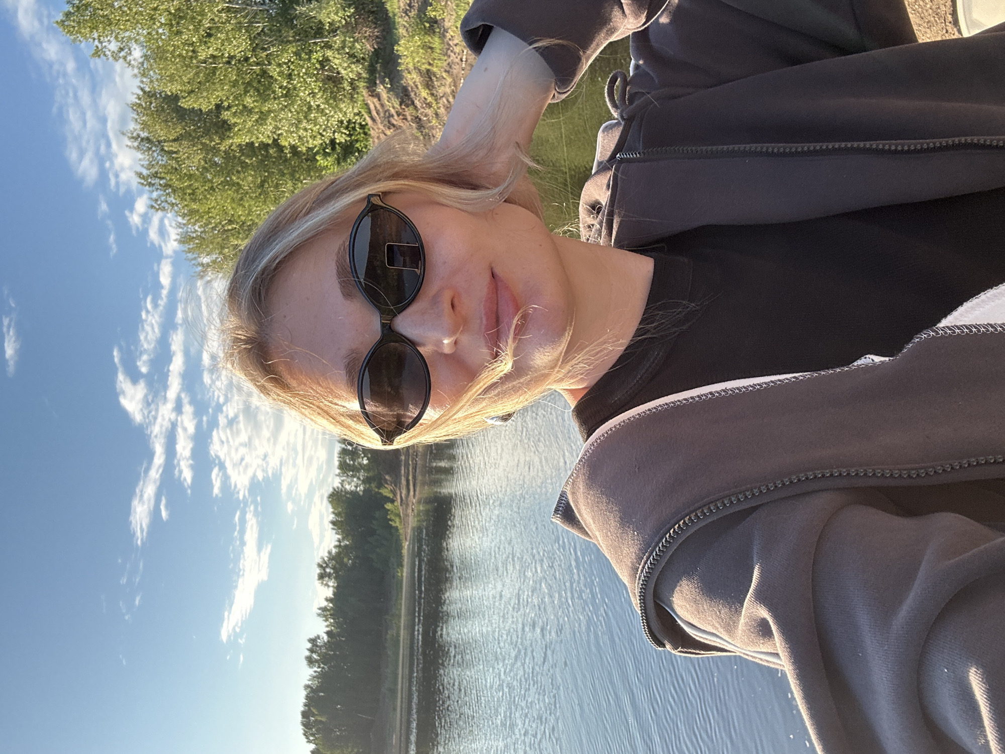 A young woman with blonde hair, wearing sunglasses and a dark hooded jacket over a black t-shirt, takes a selfie outdoors by a body of water. The background shows a calm lake or river, with a tree-lined shore and a blue sky with scattered clouds. The lighting suggests it is late afternoon or early evening. The location appears to be a natural, possibly rural or park-like setting, consistent with areas around Noviy, Russia.