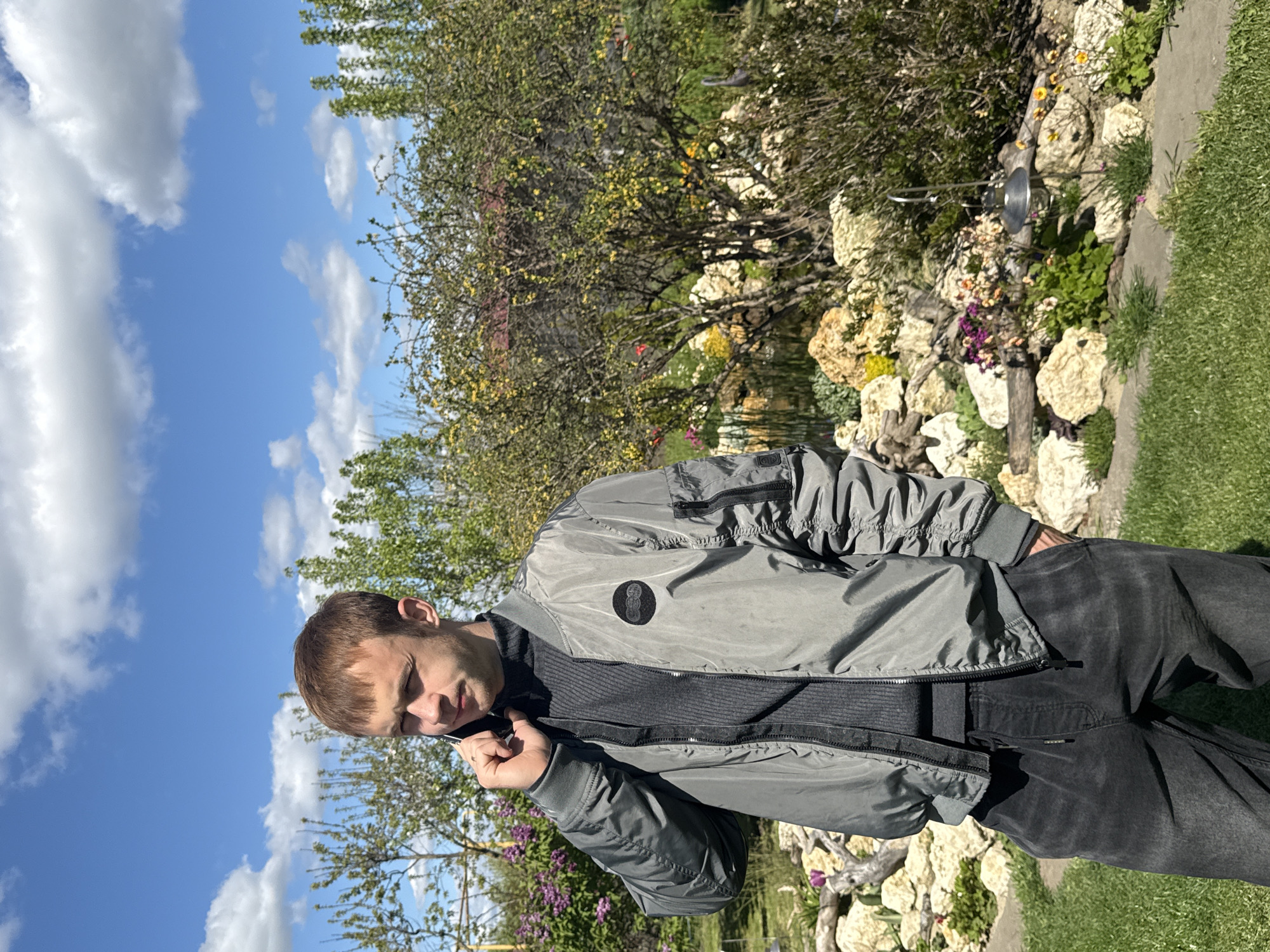 The image displays a young man standing outdoors in a sunny garden or backyard setting, likely in Pervorechenskoye, Russia. He is positioned slightly to the right of the frame, wearing a grey bomber jacket over a dark grey ribbed sweater and dark pants. His right hand is brought up towards his neck area, and his head is slightly tilted, with his eyes appearing partially closed, possibly due to the bright sunlight. He has short, light brown hair.
The background reveals a vibrant outdoor scene on a clear, bright day. The sky is a prominent blue with scattered white, fluffy clouds. To the left and behind the man are lush green trees and bushes, some with hints of yellow and reddish leaves. To his right, a decorative rock garden features various small green plants and colorful flowers in shades of purple, yellow, and orange. A section of green grass is visible in the lower right foreground. The strong, natural light suggests it is daytime. The man appears to be posing for a photograph. No visible text is discernible in the image.