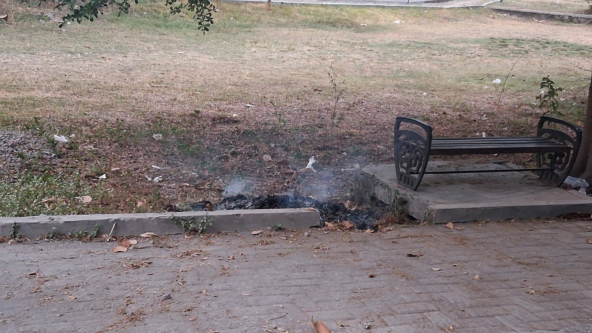 The image shows an outdoor scene with dry grass and fallen leaves covering the ground. In the foreground, there is a paved walkway made of interlocking bricks. To the left of the walkway, there is a concrete border. Behind the border, a pile of burning leaves is producing smoke. To the right of the burning leaves, a dark metal bench with decorative ends is placed on a concrete platform. The bench has wooden slats for the seat and backrest. In the background, there is more dry grass and some trees with green leaves. The overall scene suggests a park or public space with some litter and evidence of a small fire. No specific details about the city of Yogyakarta, Indonesia, are visible in the image.