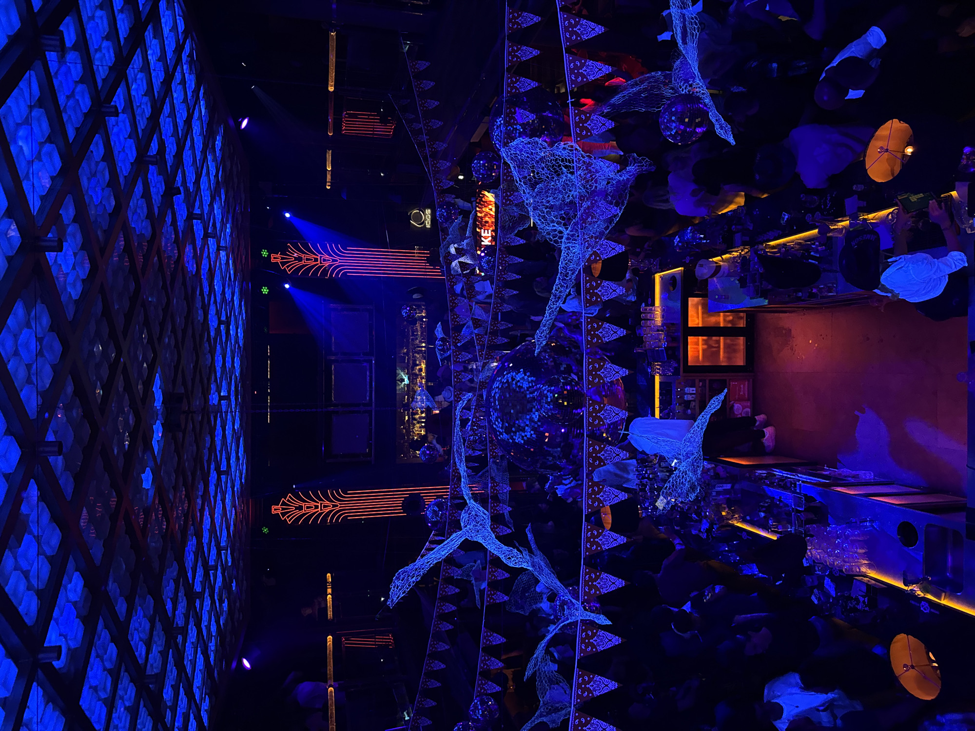 The image captures a bar or club interior in Sofia, Bulgaria. The lighting is predominantly blue, with orange accents. The left wall is covered in a blue geometric pattern. Hanging sculptures resembling birds or stylized animal figures are suspended from the ceiling or fixtures. Mirrored elements are present, reflecting the bar and people. People are present, clustered around the bar, suggesting an active social scene. The bar itself is illuminated with orange or yellow lights highlighting the bar area, the bottles, and glasses.