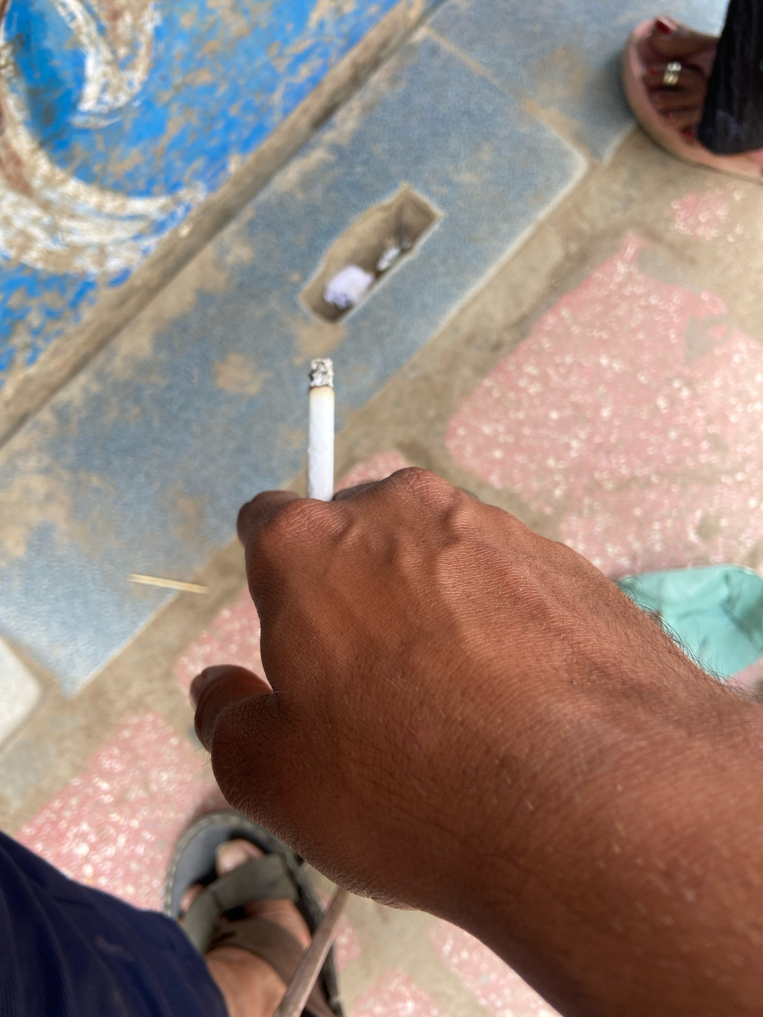 An extreme close-up shot focuses on a person's hand holding a lit cigarette. The hand, with visible veins and hair, is positioned in the foreground. The background suggests an outdoor environment with a tiled floor featuring blue and pink patterns. A small, rectangular opening in the blue tiled area contains what appears to be a crumpled piece of white paper. A toothpick lies near the cigarette. In the upper right corner, a person's foot wearing a sandal and a ring on their toe is partially visible. The overall scene implies a casual setting, potentially a public space in Indore, India.