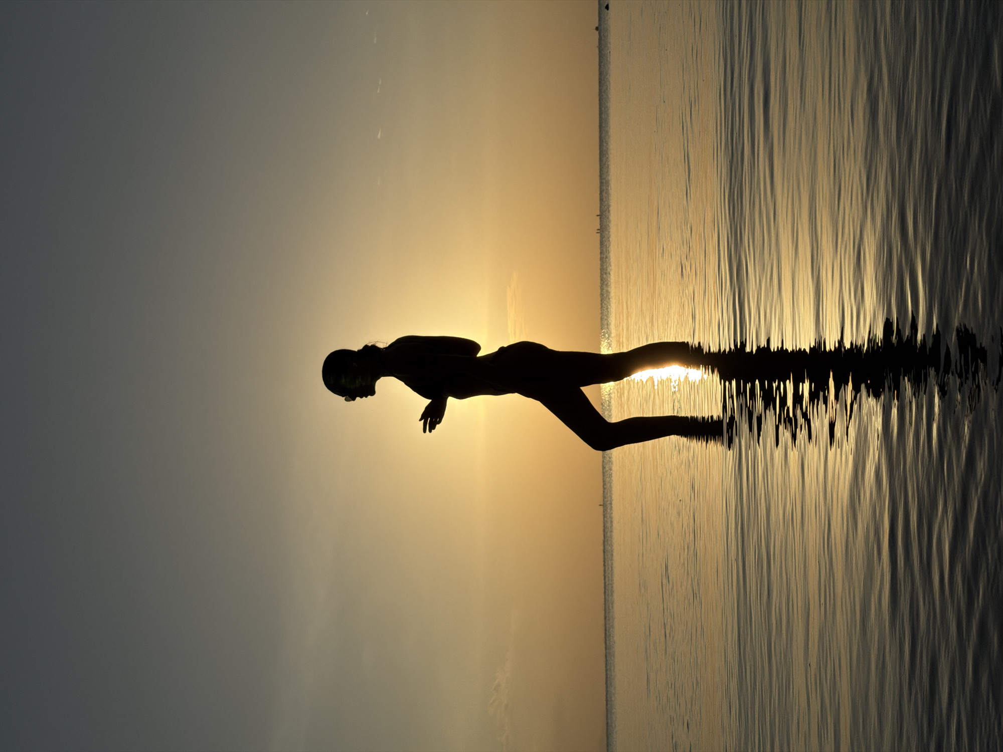 A silhouette of a person wearing sunglasses stands in shallow water against a sunset. The person is facing to the left, with their right arm bent at the elbow and their left arm extended slightly forward. The sun is setting in the background, casting a warm orange glow across the sky and reflecting on the water. The water is calm, with gentle ripples. The location is Ko Samui District, Thailand, during sunset.