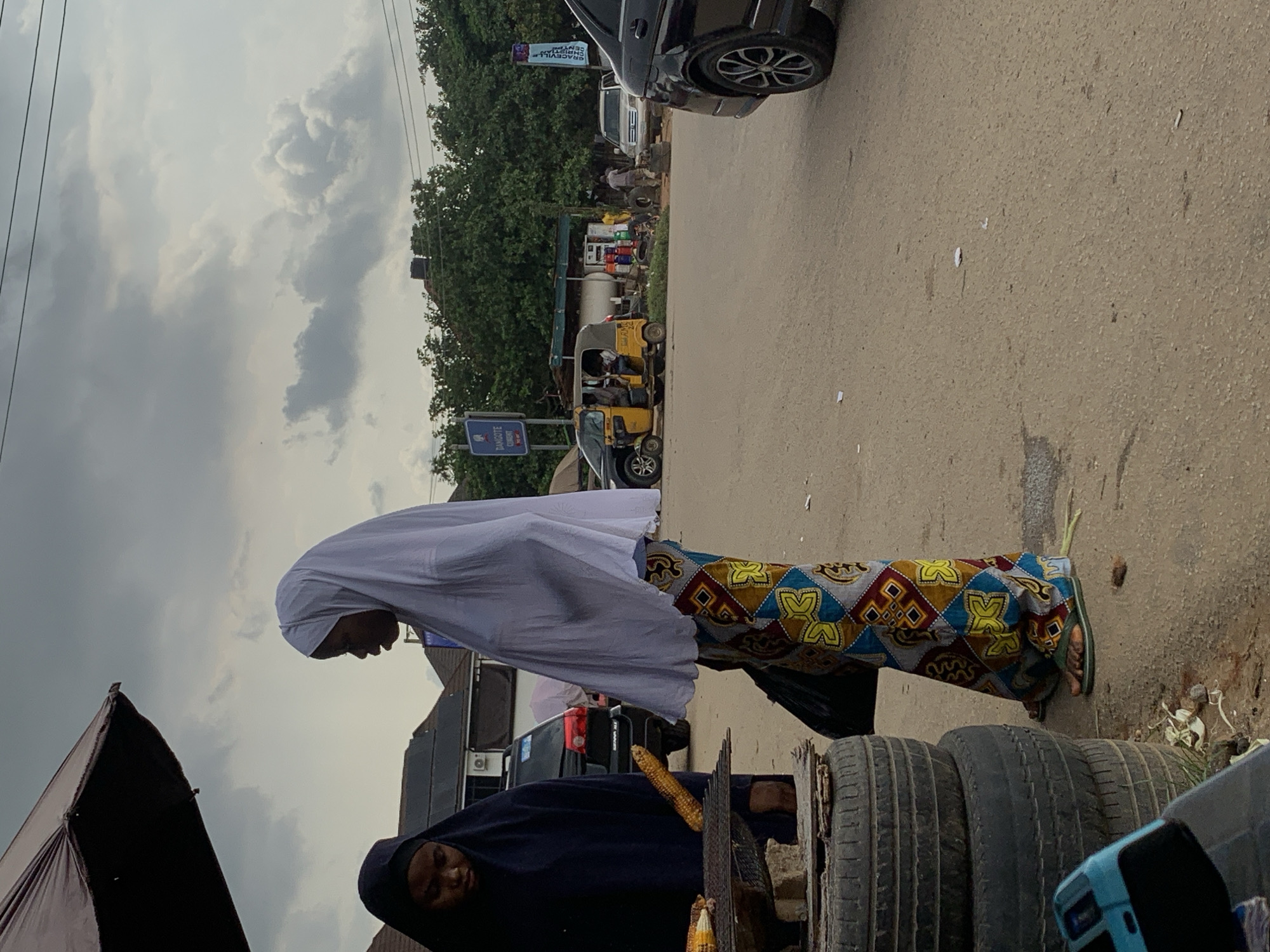 In Abuja, Nigeria, an outdoor scene depicts two women near a roadside food stall under a large, dark umbrella. The woman on the left, dressed in a dark hijab, is partially visible, tending to corn roasting on a metal grate. The woman on the right wears a white hijab and a vibrantly patterned skirt. Stacked tires and other debris are visible on the ground. In the background, a yellow auto-rickshaw, several cars, and a small building with a fuel station sign are present, suggesting a commercial or transport hub. The sky is overcast with dramatic clouds.