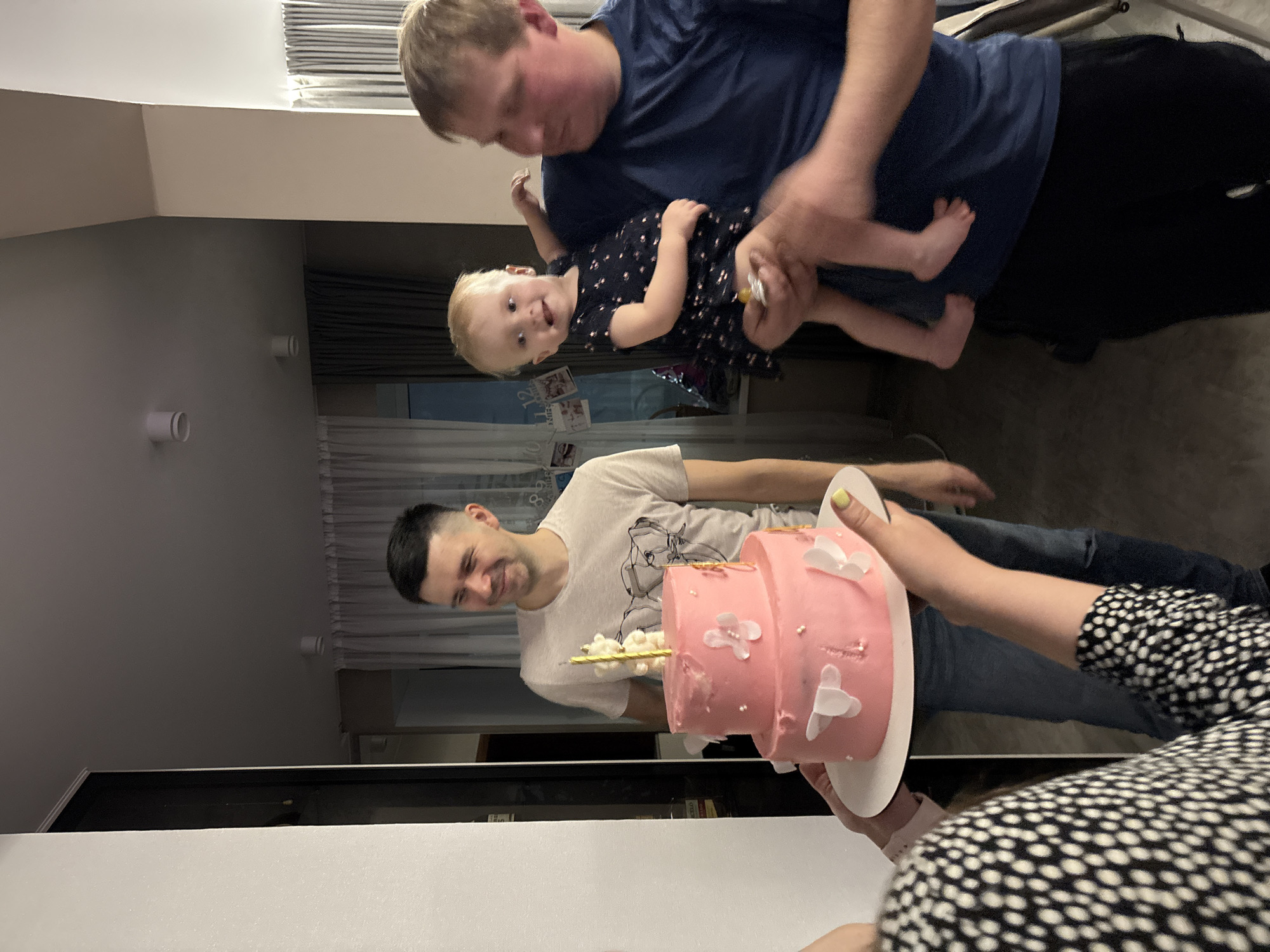 The image depicts an indoor birthday celebration scene, likely in a residential setting. A fair-skinned man in a white t-shirt stands in the middle, smiling towards a pink, two-tiered cake adorned with white butterfly decorations, which is being held by a person wearing a black and white polka-dot patterned shirt. To the upper right, a second fair-skinned man, wearing a blue t-shirt, is holding a blonde toddler dressed in a dark, dotted dress. The backdrop shows a window with blinds and some photographs or cards. The room has a modern feel with light walls and ceiling. There is not enough information to specifically say that the location is in Garevaya, Russia.