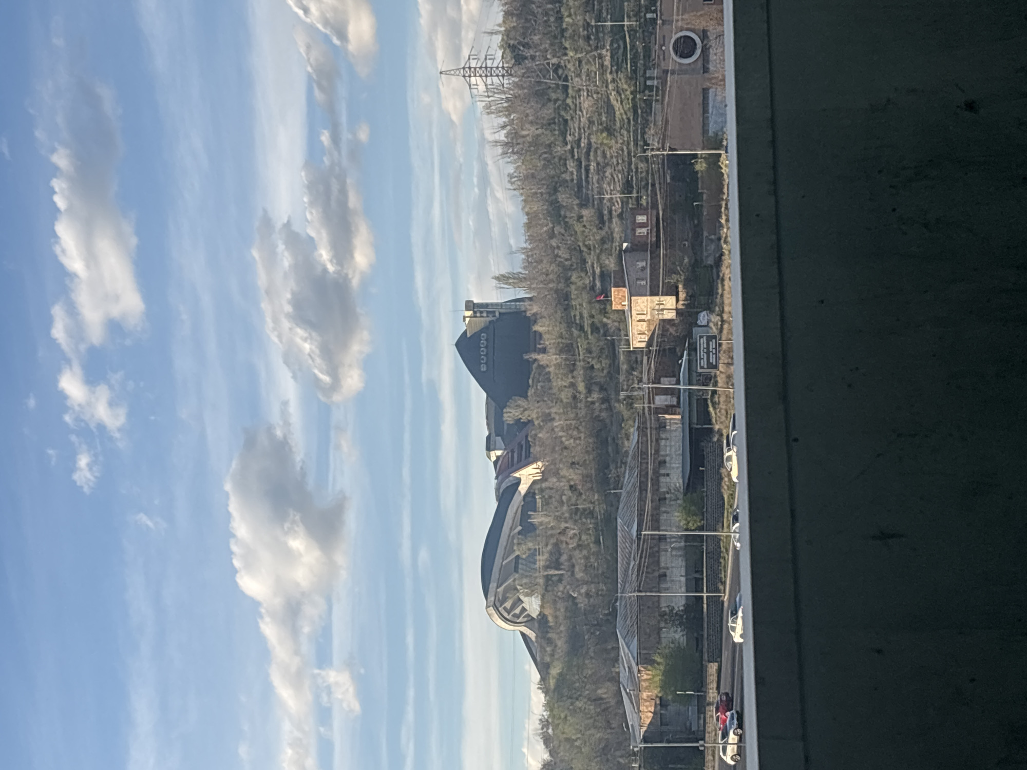 The image captures a view of Yerevan, Armenia, through a frame, likely from a bridge or elevated structure. Prominently featured in the landscape are:
* Karen Demirchyan Sports and Concerts Complex: This large, pyramid-shaped building with the word "DUQOD" on its face is a central landmark.
* Tsitsernakaberd Armenian Genocide Memorial: The distinctive, curved structure of this memorial is also visible.
* Environmental Details: Trees, vegetation, and other buildings create a dense urban landscape.
* Sky and Clouds: A blue sky with scattered white clouds forms the backdrop.
* Power Lines: A utility tower is visible in the distance.
* Transportation: A street runs along the right side of the frame, with cars parked or moving along it.
* Concrete Structure: The photograph is taken from within or behind a large concrete structure, obscuring part of the view.