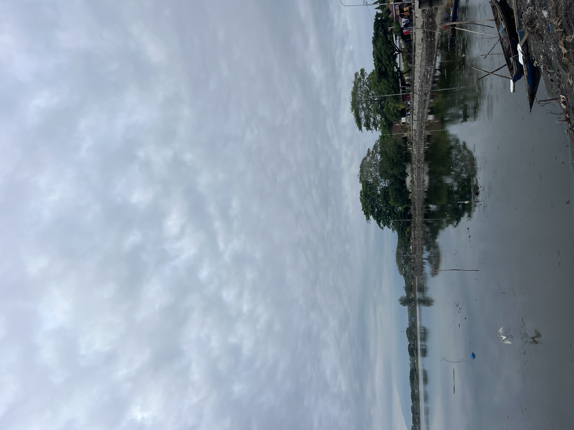The image depicts a cloudy, overcast scene near a body of water in Lhokseumawe, Indonesia. A narrow bank separates the water from an area with trees and some structures. Debris and litter are visible in the water and along the edge. A small boat is visible on the right edge. The water reflects the sky and the trees. The atmosphere is dull and gray.