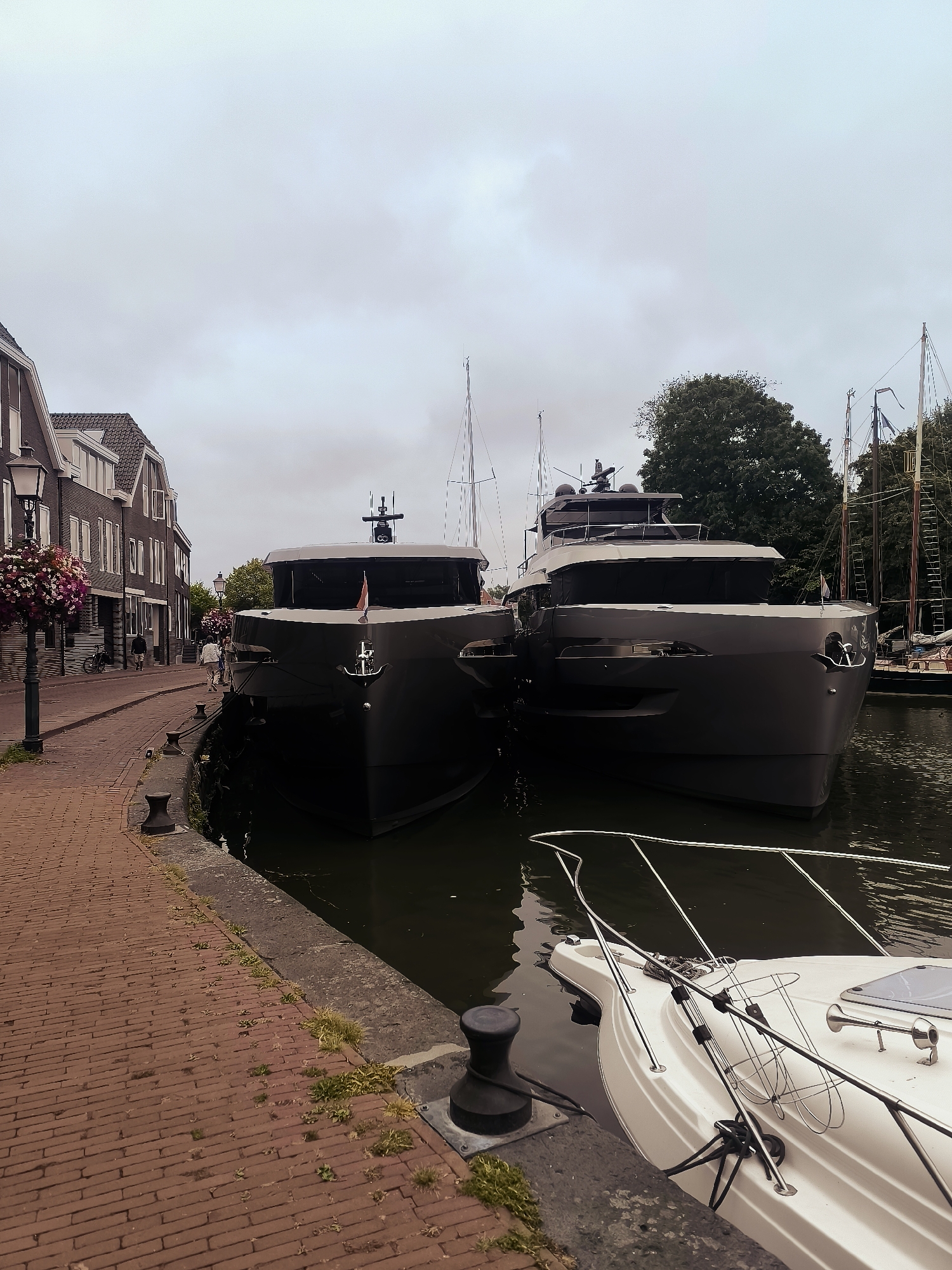 Two large, dark-colored yachts are docked side-by-side in a canal in Hoorn, Netherlands. The yachts have sleek, modern designs with sharp lines and large windows. The foreground shows the bow of a white speedboat also docked in the canal. To the left of the canal is a brick walkway and a street lined with historic brick buildings. There are street lamps with flower baskets hanging from them. A few people are walking on the street and along the canal. In the background, trees and masts of sailboats are visible against a cloudy sky. The scene appears to be a picturesque canal view in a European city.