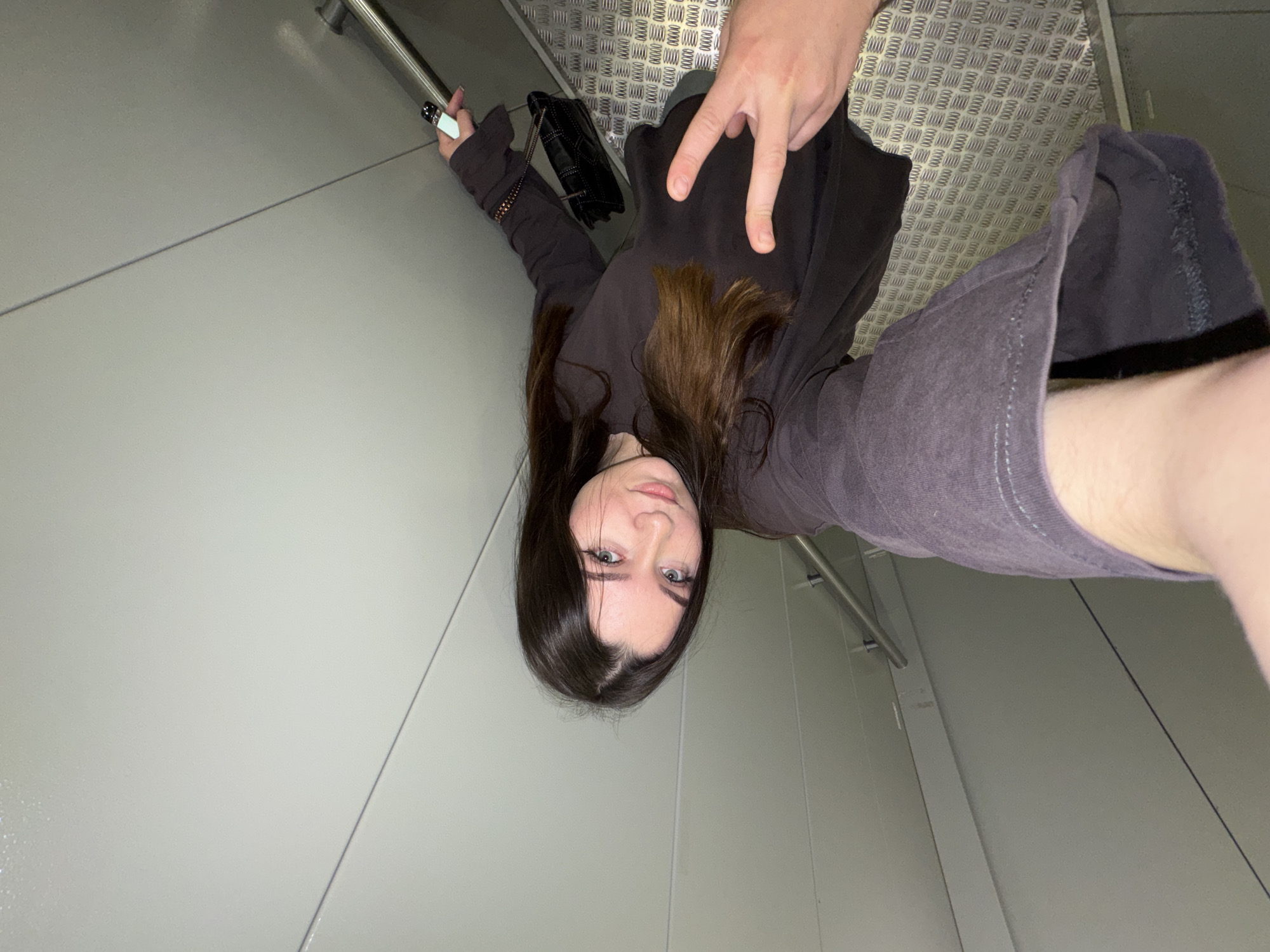 A young woman with long, dark brown hair and blue eyes is taking a selfie on a metallic staircase. She is wearing a dark gray long-sleeved shirt. Her left arm is extended, holding the camera, and her right hand is near her chest, forming a peace sign. A black quilted handbag with a chain strap is visible on her right side. The staircase has a textured metal floor and a silver railing. The walls are a light gray color, with visible seams. The scene appears to be indoors, possibly in a modern building or public transport. The specific location in Shirkovo, Russia, is not identifiable from the visual information in the image.