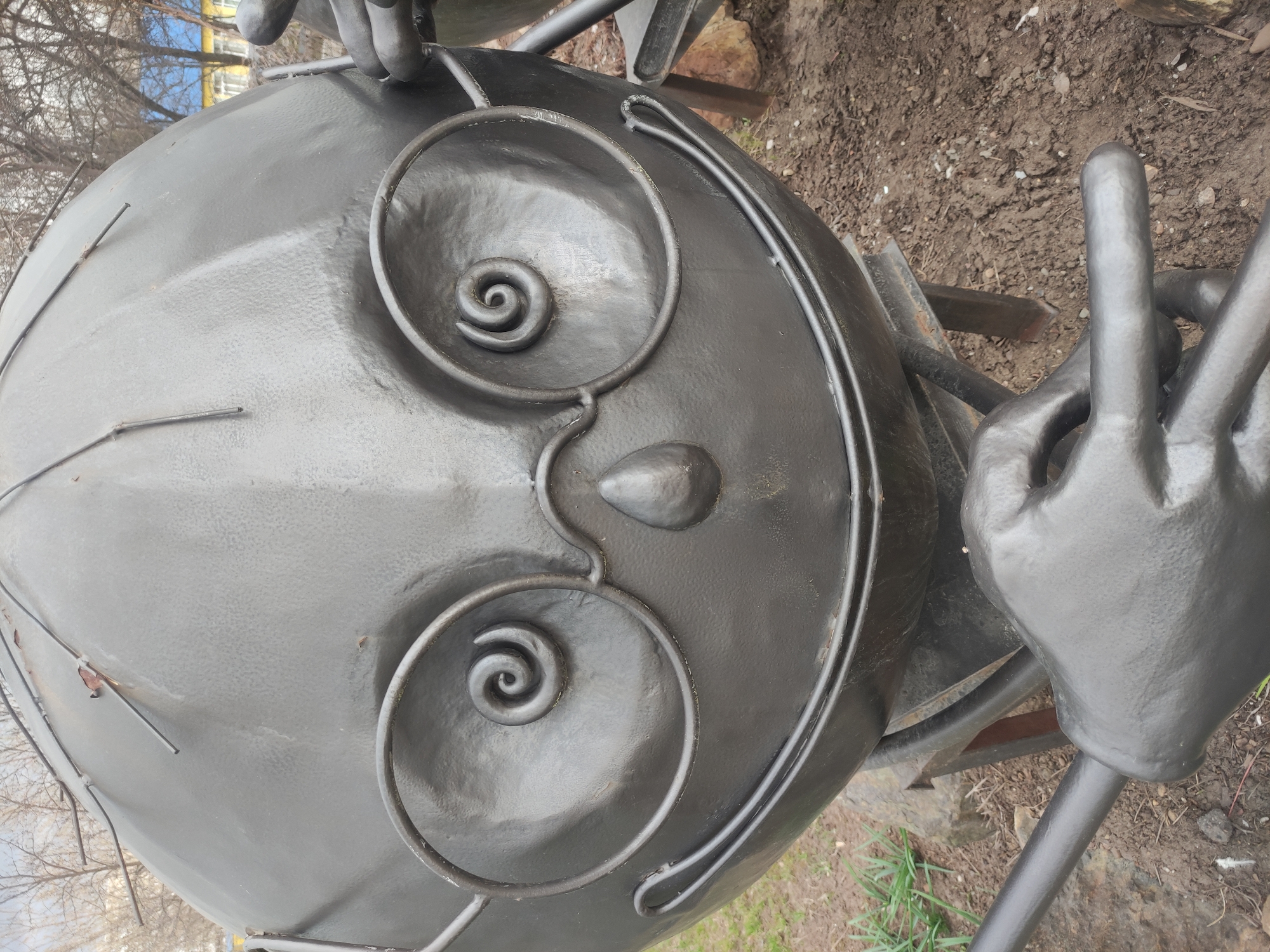 The image shows a close-up of a metal sculpture of a face. The face is round, with spiral shapes for eyes and a raised, conical nose. A thin, curved metal bar runs around the perimeter of the face. To the right of the face is a metal hand sculpture. The background consists of dirt and sparse vegetation, suggesting the sculpture is located outdoors. The location is given as Unknown, Unknown.