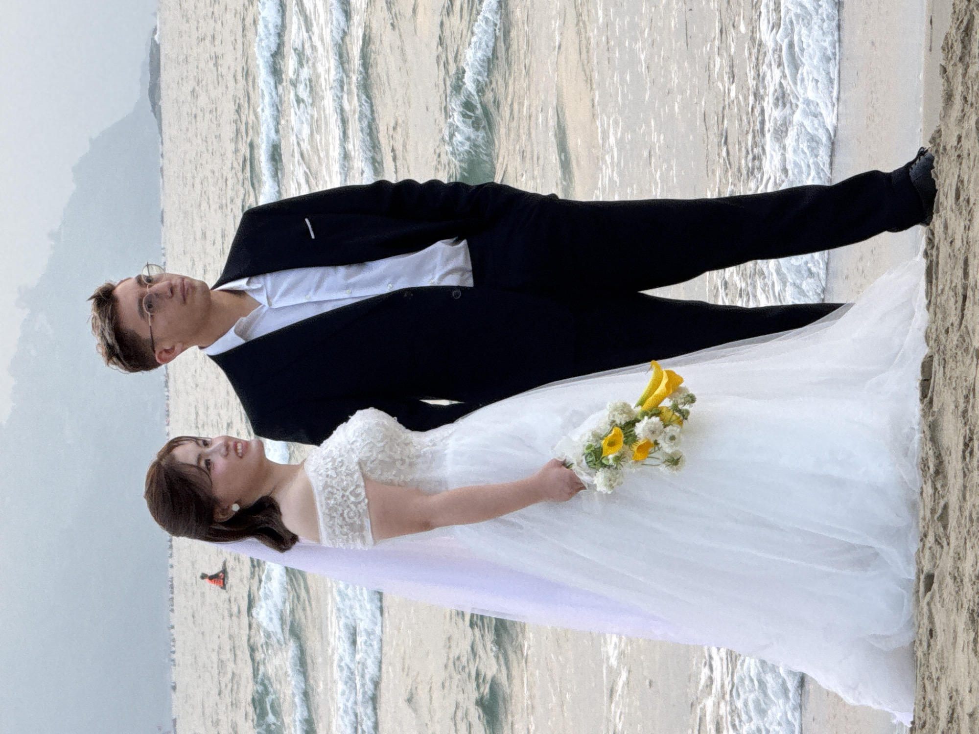 A wedding photo captured on a beach in Đà Nẵng, Vietnam shows a Caucasian man and Asian woman. The man, dressed in a black suit with a white shirt, wears glasses. The woman is in a white wedding gown holding a bouquet of yellow and white flowers. The background features a beach with gentle waves and a distant mountain or island. A red object can be seen in the sea in the background.