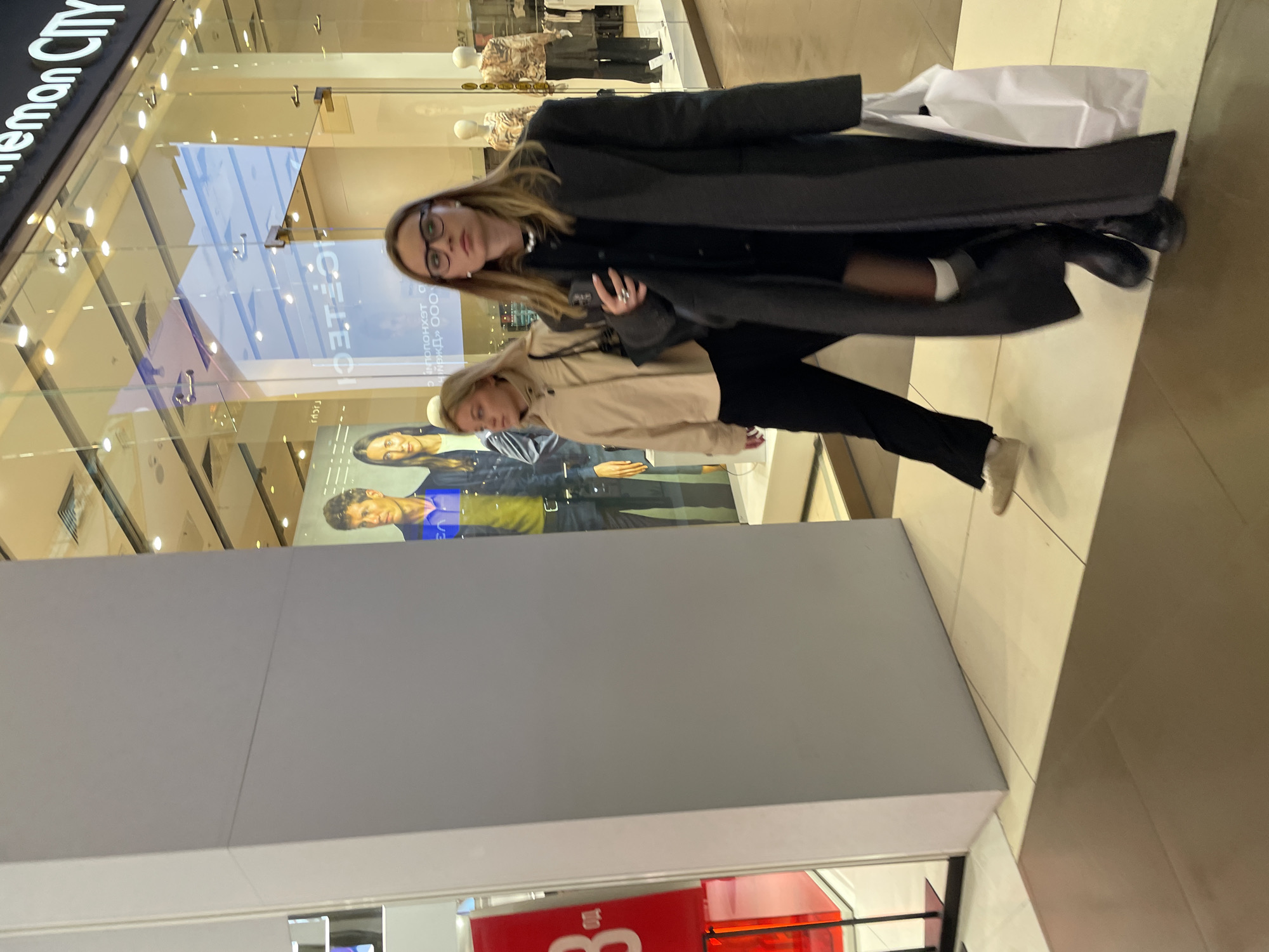 Two women pose in front of a shop window in a mall. The woman in the foreground wears glasses, a black coat, a black dress with white stockings, and black shoes. She holds a black phone in her hand and a white shopping bag rests near her feet. The woman behind her has blonde hair and wears a beige jacket and black pants with white sneakers. The shop window behind them displays an advertisement featuring two men. The image is taken indoors in a mall, with a section of the mall's ceiling and structure visible, including the "man City" sign. A storefront with red signage is partially visible at the bottom of the frame. The location is in a shopping mall, the city of Saint Petersburg, Russia.