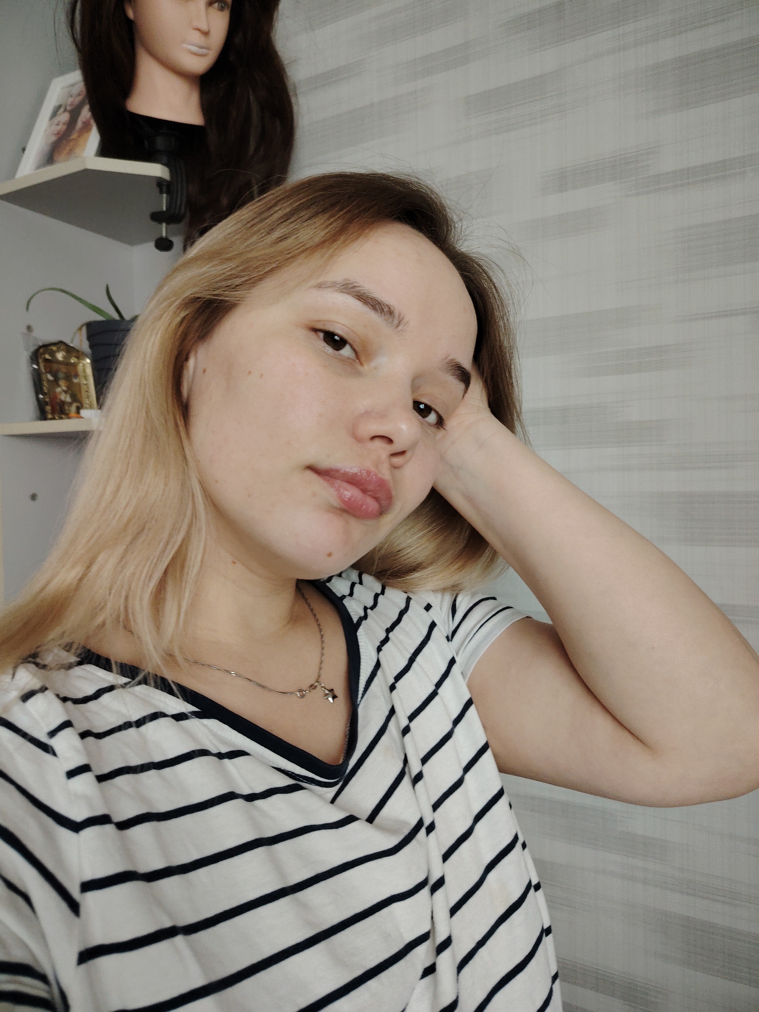 A woman is posing for a selfie in an indoor setting in what appears to be a residential space in Марковский, Russia. She has light blonde hair with darker roots, light skin, and is wearing a black and white striped shirt. She is resting her head on her right hand, and a silver necklace with a star charm is visible around her neck.
In the background, a shelf is visible with a mannequin head with dark brown hair, a framed picture, a small plant, and a decorative golden object. The wall behind the woman is covered with a light-colored wallpaper with horizontal striped texture. The lighting appears to be soft and natural.