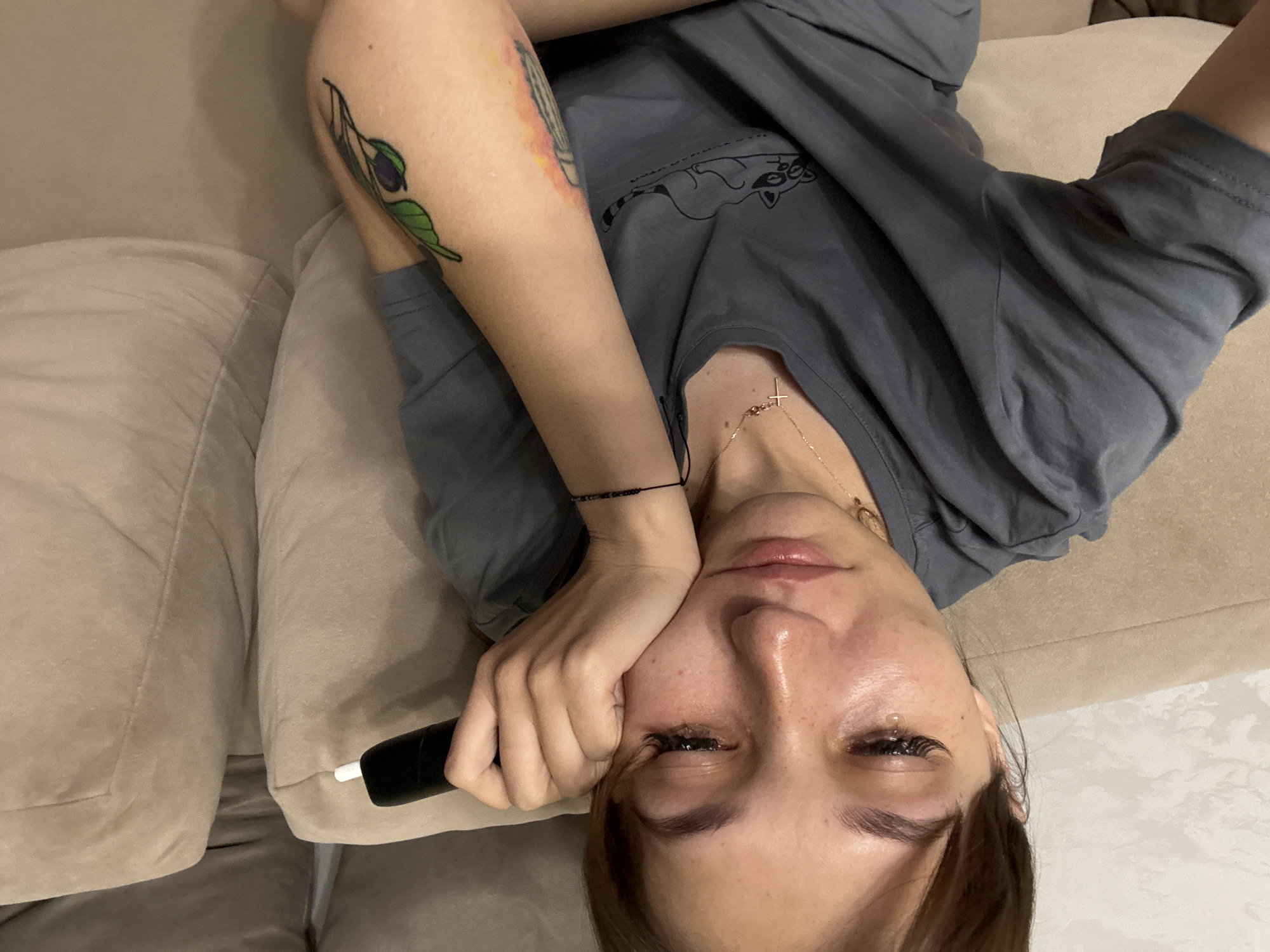 A young woman is sitting on a beige sofa, holding a black and white electronic device to her cheek. Her face is tilted downwards, and her eyes are closed, with tears visible on her cheeks and eyelashes. She has on a gray t-shirt with a drawing of a raccoon. She is also wearing a delicate gold necklace with a cross pendant and a beaded bracelet on her left wrist. Tattoos are visible on her right arm. The background shows the textured fabric of the sofa and a hint of a light-colored wall. The scene appears to be indoors, likely a living room. The location of Novokuznetsk, Russia, is not visually represented in the image.