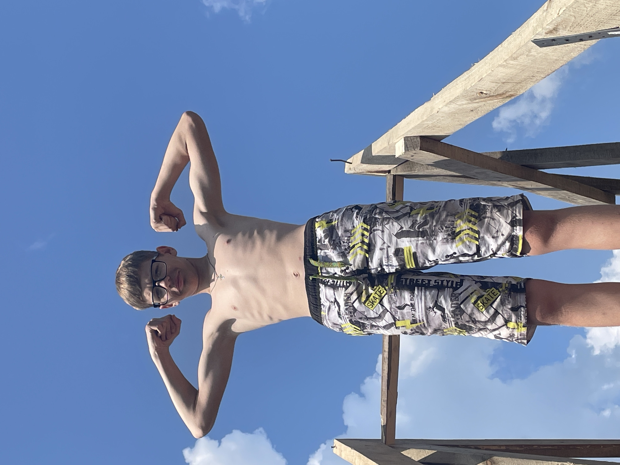 A young man is posing with his arms flexed against a clear blue sky with scattered white clouds. He is wearing black-rimmed glasses and patterned swim trunks with yellow and black designs. The swim trunks have text that includes "SKATE BOARD," "STREET STYLE," and "I'M THE BOX." He is standing in front of a wooden structure, possibly under construction. The image is oriented vertically, with the subject appearing upside down relative to the horizon. The lighting suggests it is daytime, with a bright sun casting shadows. The overall impression is one of youthful confidence and a casual outdoor setting.