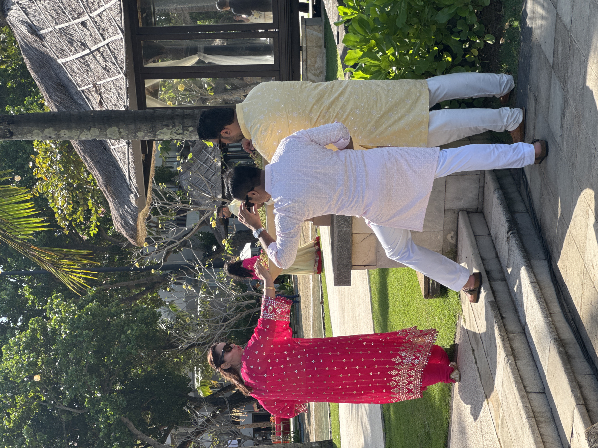 The image depicts three people outdoors in Kuta Selatan, Indonesia. A woman in a long-sleeved pink and gold embroidered garment stands in the foreground. Two men stand behind her; the first is wearing a white Kurta and pants and the second is wearing a yellow Kurta and pants. The men appear to be posing for a picture. The background includes trees, a building with a thatched roof, and greenery. The setting appears to be a resort or outdoor venue in Kuta Selatan.