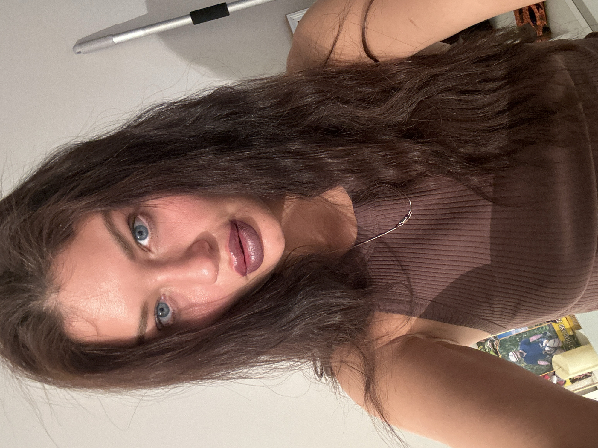 A young woman is posing for a selfie. She has long, wavy brown hair, fair skin, and blue eyes. She is wearing a brown ribbed tank top and a thin silver necklace. Her lips are coated in dark, glossy lipstick. The background is blurry and indistinct, but hints of a white wall, a silver rod with a black handle, and a partially visible object with a Thailand label can be observed. The photo appears to be taken indoors. There is no verifiable evidence to determine whether this image was taken in Khimki, Russia.