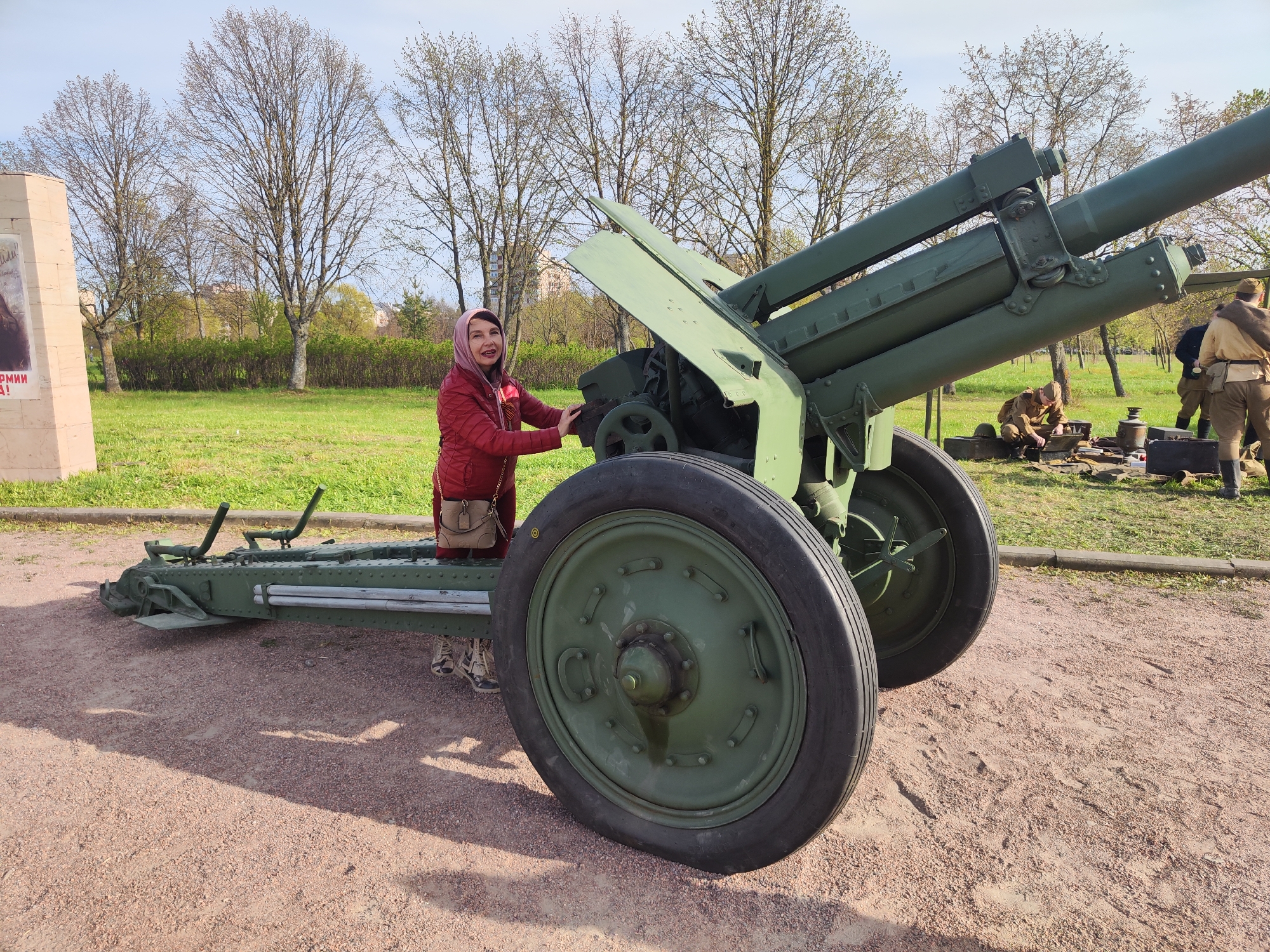 A woman in a red jacket and pink headscarf is standing next to a green artillery gun in a park setting. The gun is on a gravel area with a grassy lawn and trees in the background. To the right of the gun, two men in military-style uniforms are gathered around a box. A monument stands to the left of the woman.