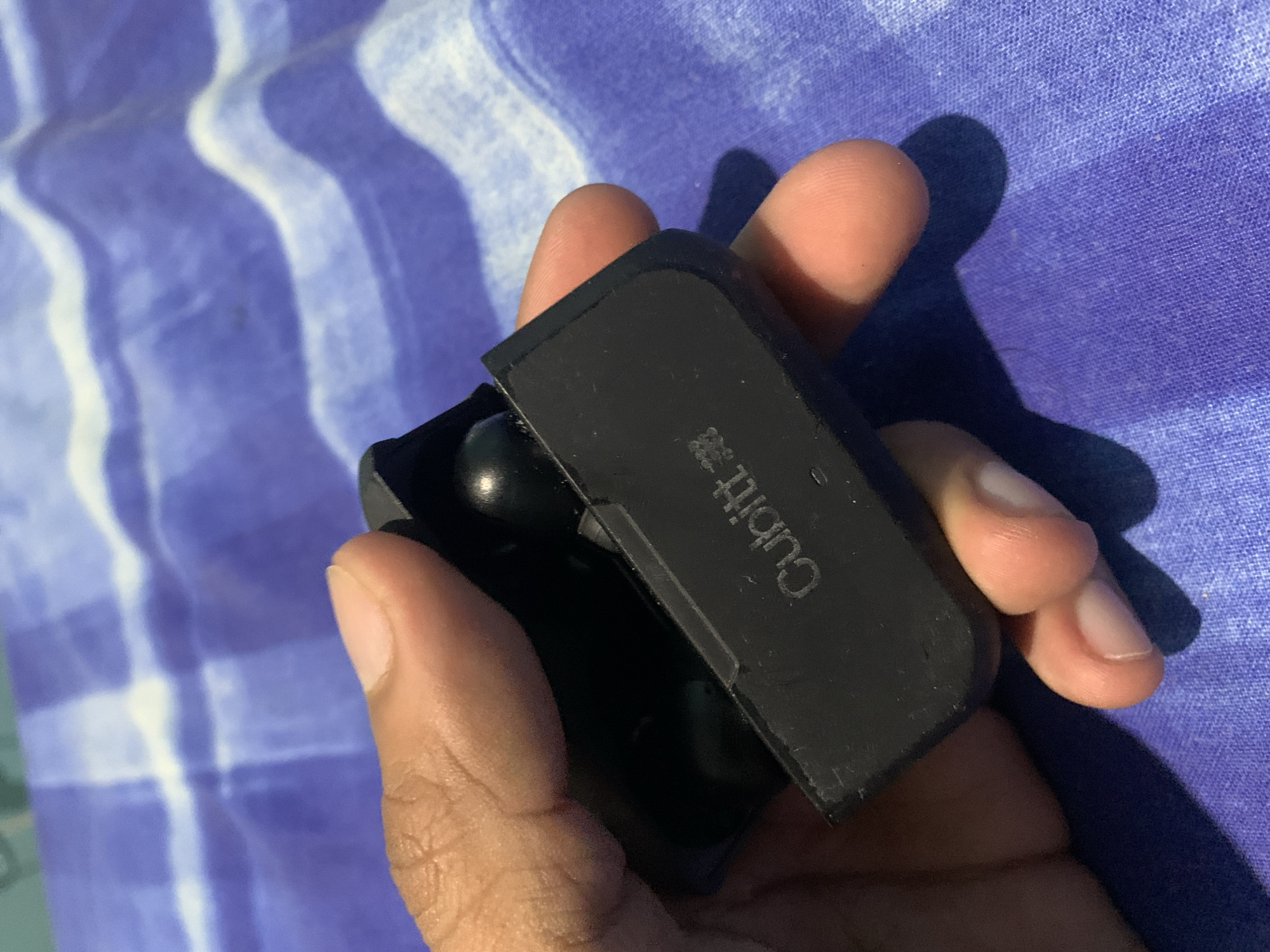 A human hand is holding a black, open case with "Cubitt" printed on the lid. Inside the case, two black earbuds are visible. The background is a patterned fabric with blue and white stripes. The location in Santa Ana, Venezuela is not discernible from the visual information provided.